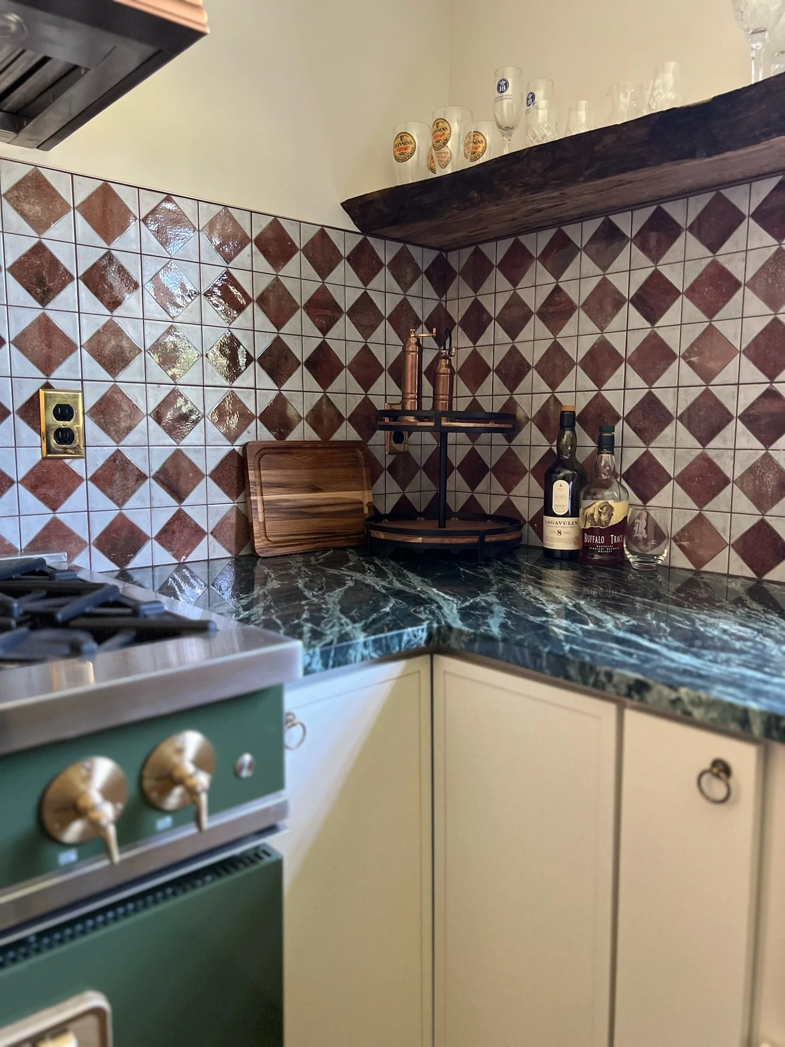 Kitchen corner with a green marble countertop, beige cabinets, a backsplash of diamond-shaped brown and beige tiles, and a wooden shelf with glassware. There are bottles of alcohol and a wooden cutting board on the counter.
