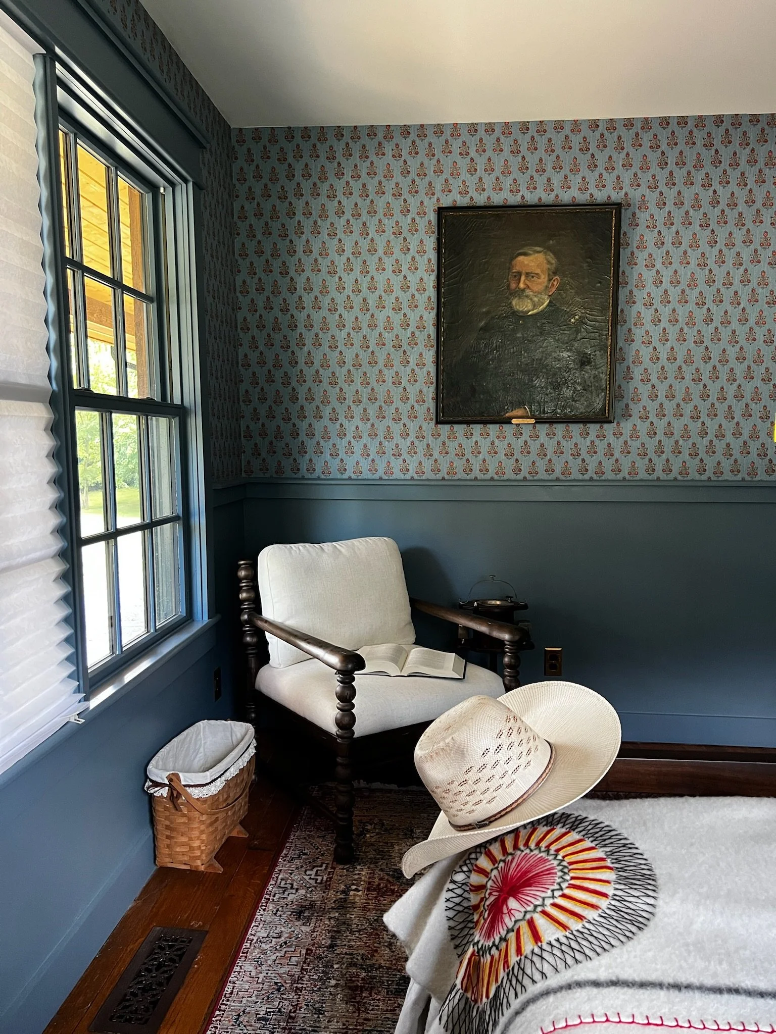 A cozy corner of a room with blue wallpaper and wooden furniture. There is a window with white blinds, a bench with a book on it, a sunhat, a cowboy hat on a table, a framed portrait of a man with a beard on the wall, and a small speaker on a side ta