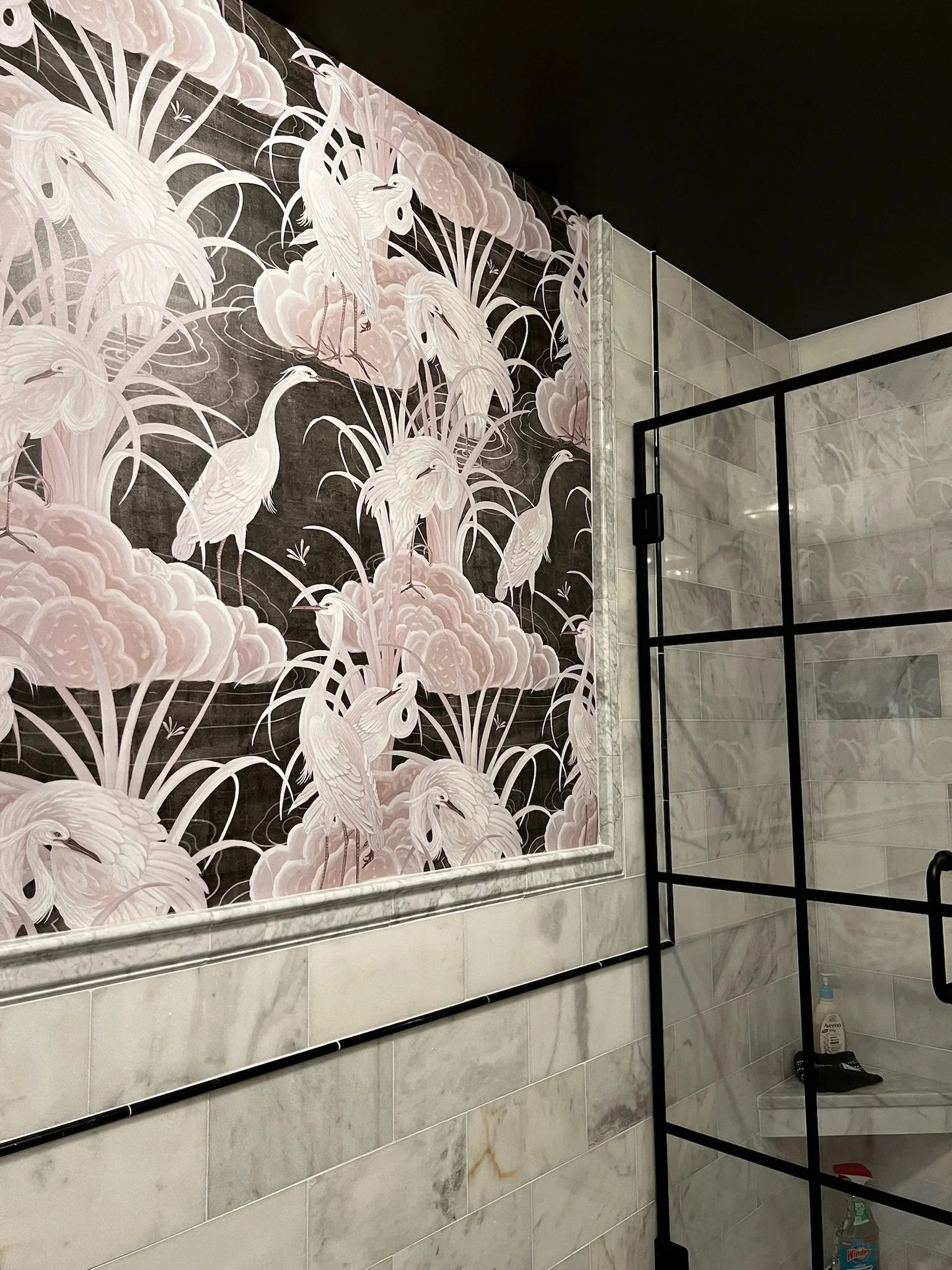 Decorative wall featuring a mural with white herons and pink clouds, located above a marble-tiled wall in a bathroom. Black metal frame of a shower enclosure is visible.
