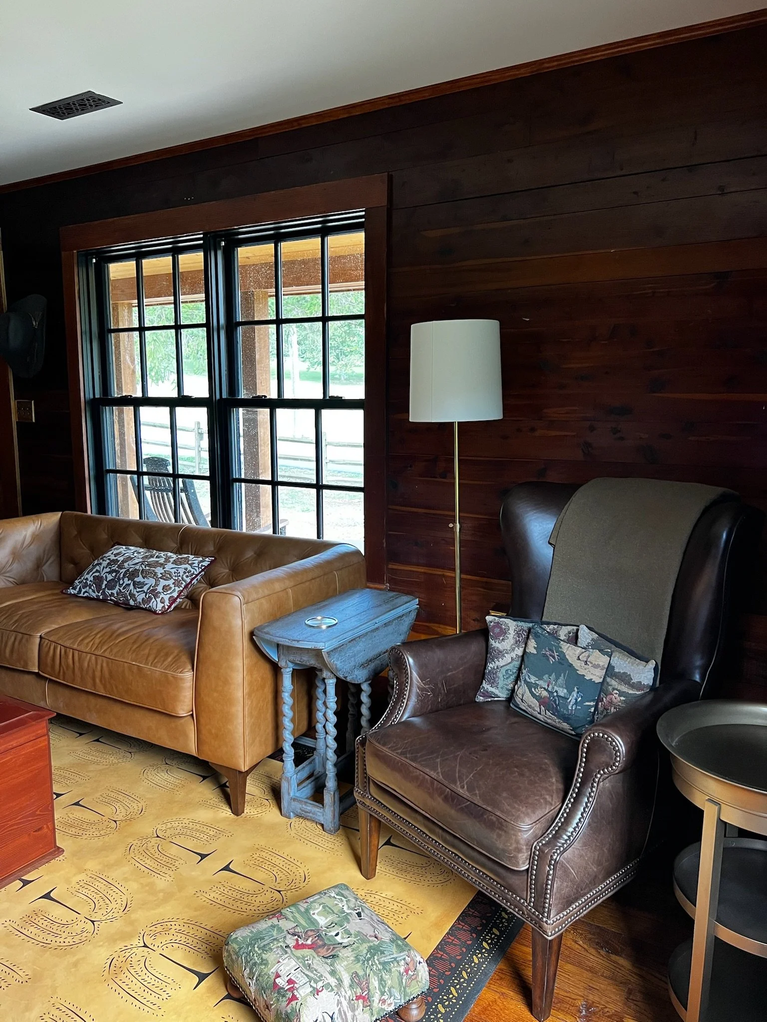 Living room with a large window, tan leather sofa, brown leather armchair with decorative pillows, small blue side table, tall white floor lamp, and a yellow carpet with a floral print.