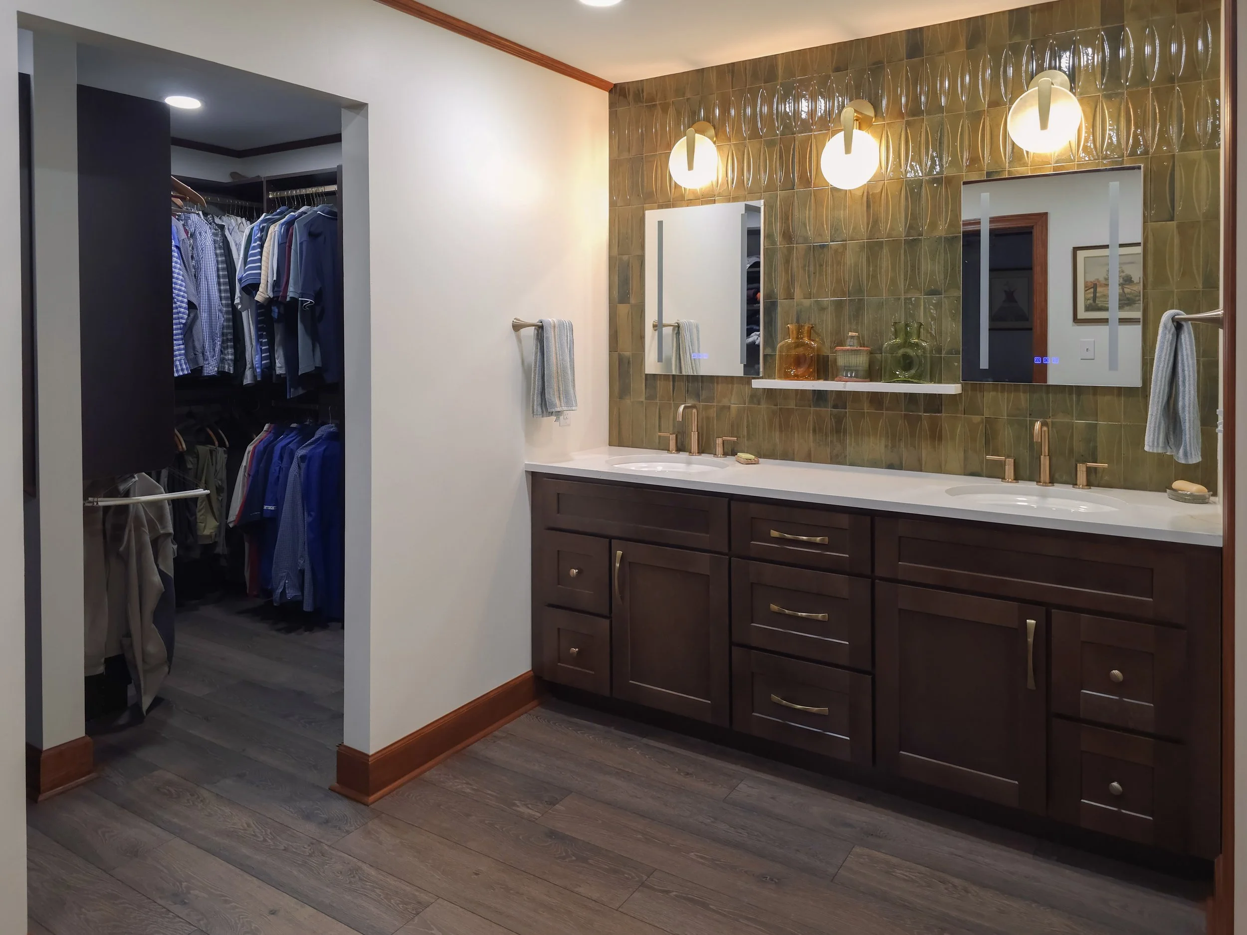 Modern bathroom with double vanity, dark wood cabinets, white countertop, two mirrors, gold fixtures, decorative lighting, and a walk-in closet with clothing.