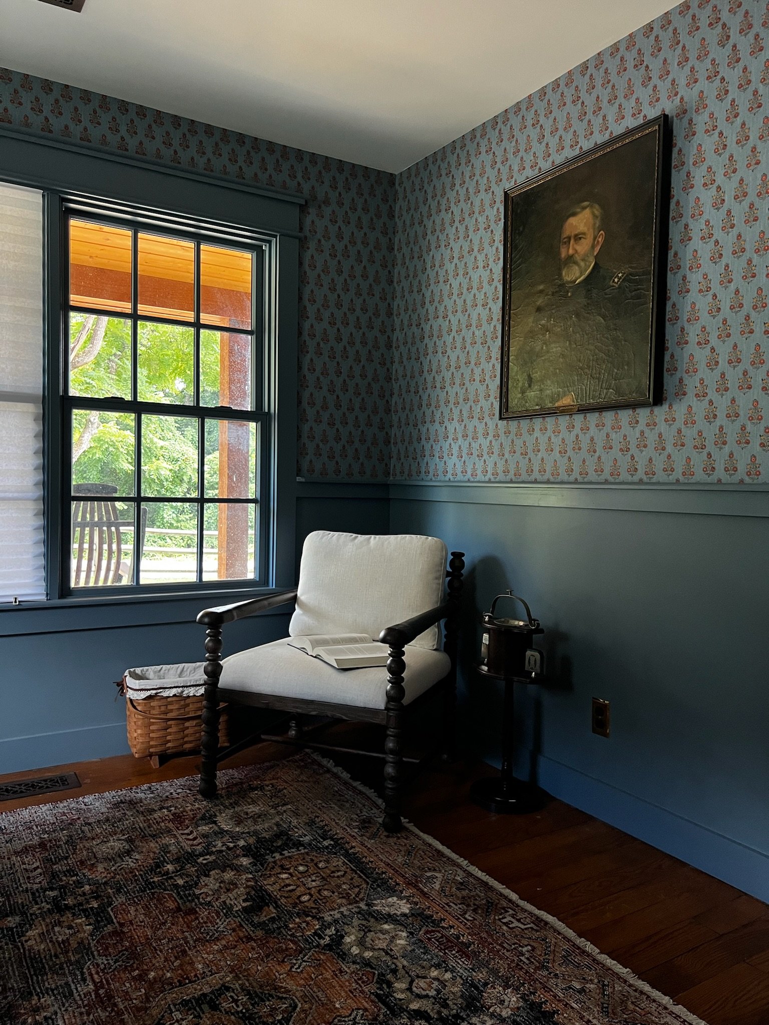A cozy corner in a blue-themed room with a window showing greenery outside, a white cushioned wooden armchair with an open book on it, a basket on the floor, a patterned area rug, and a portrait of a man in military uniform on the wall.