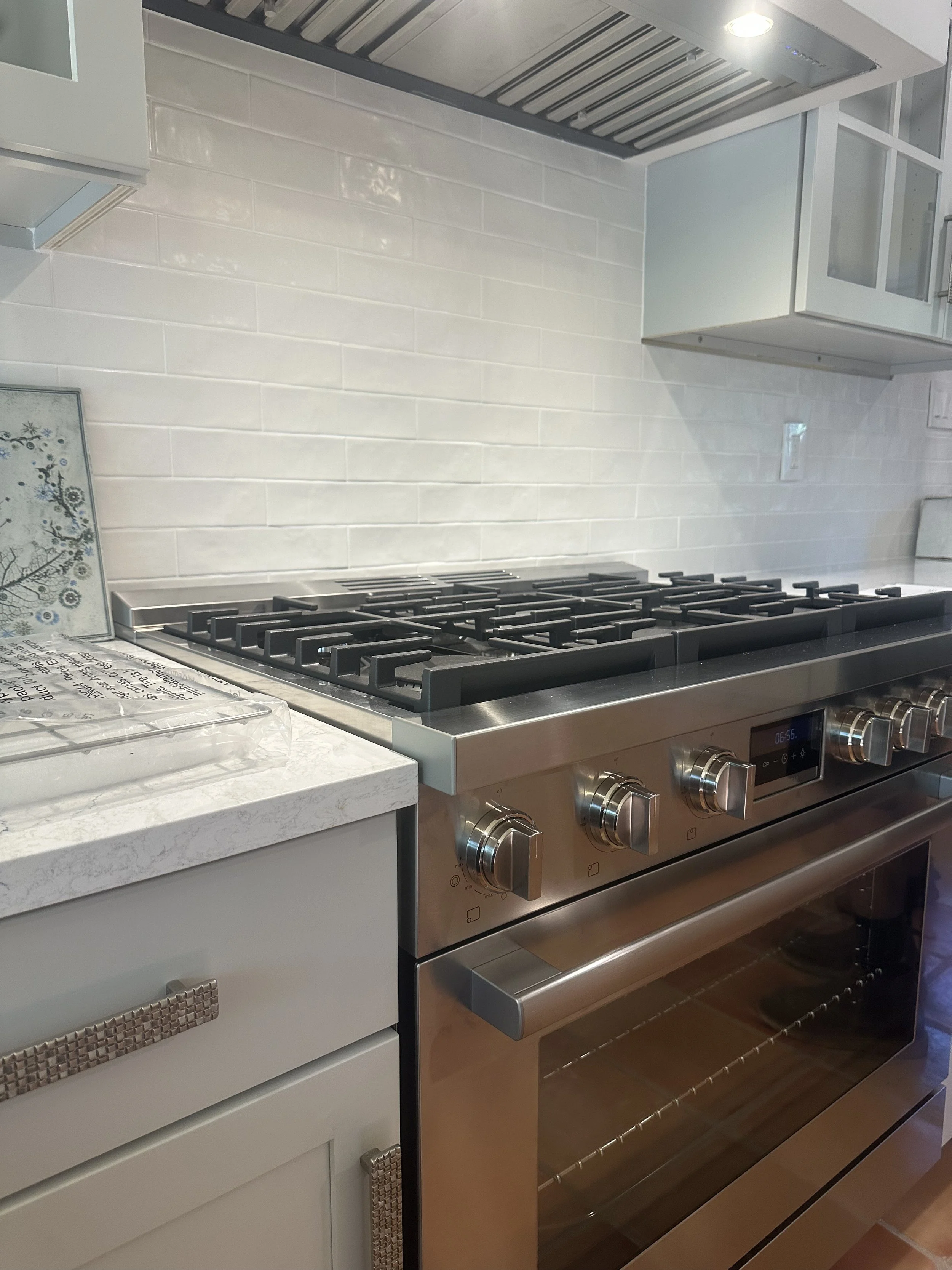 Stainless steel gas stove with five burners and a digital clock, white marble countertop, white cabinet with textured handle, white tiled backsplash, and overhead range hood in a modern kitchen.