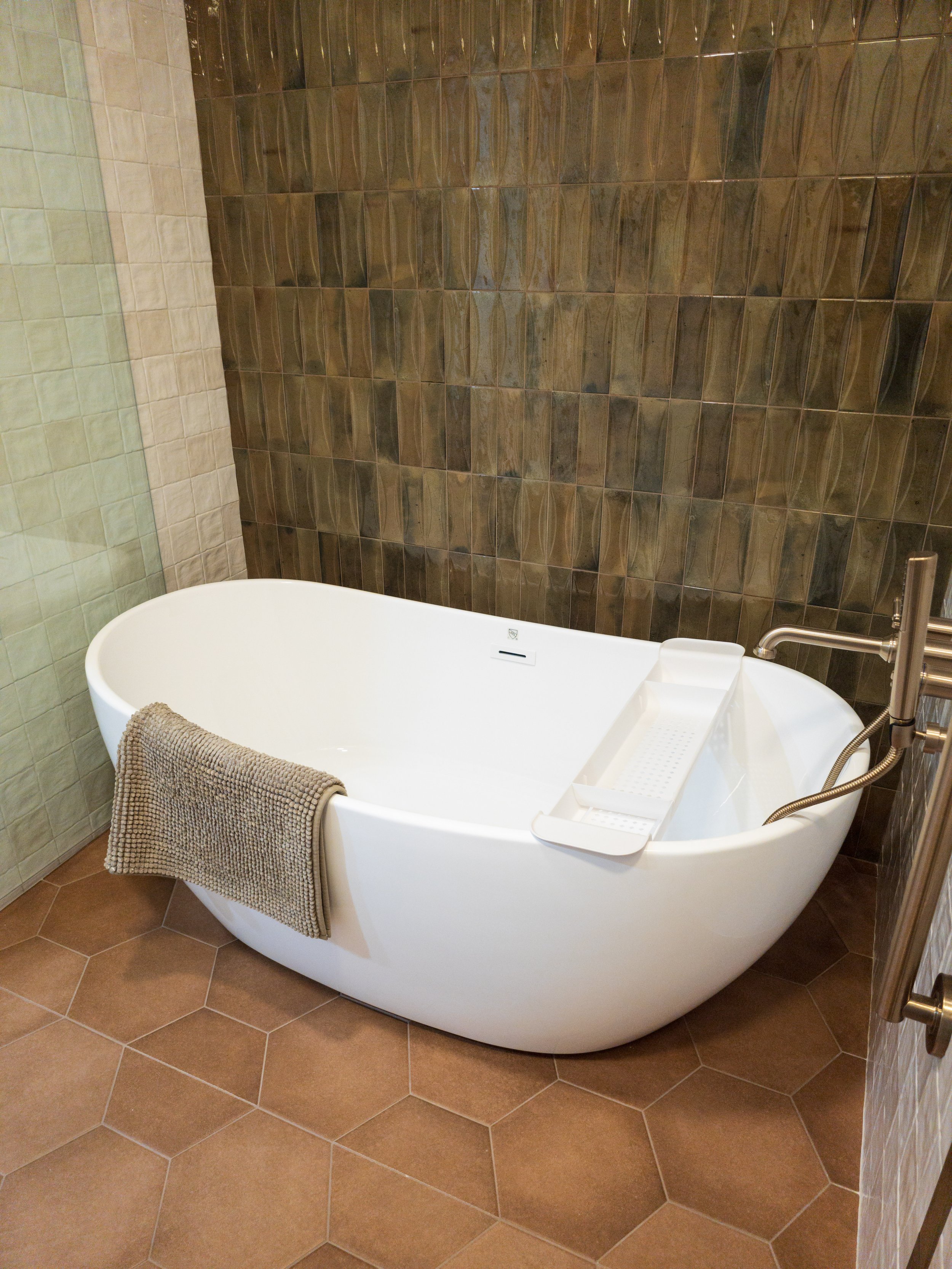 Modern freestanding white bathtub with a towel hanging on the side, positioned on a tiled bathroom floor with brown hexagonal tiles. The background features a contrast of two tile walls—one with greenish tiles on the left and dark brown tiles on the 