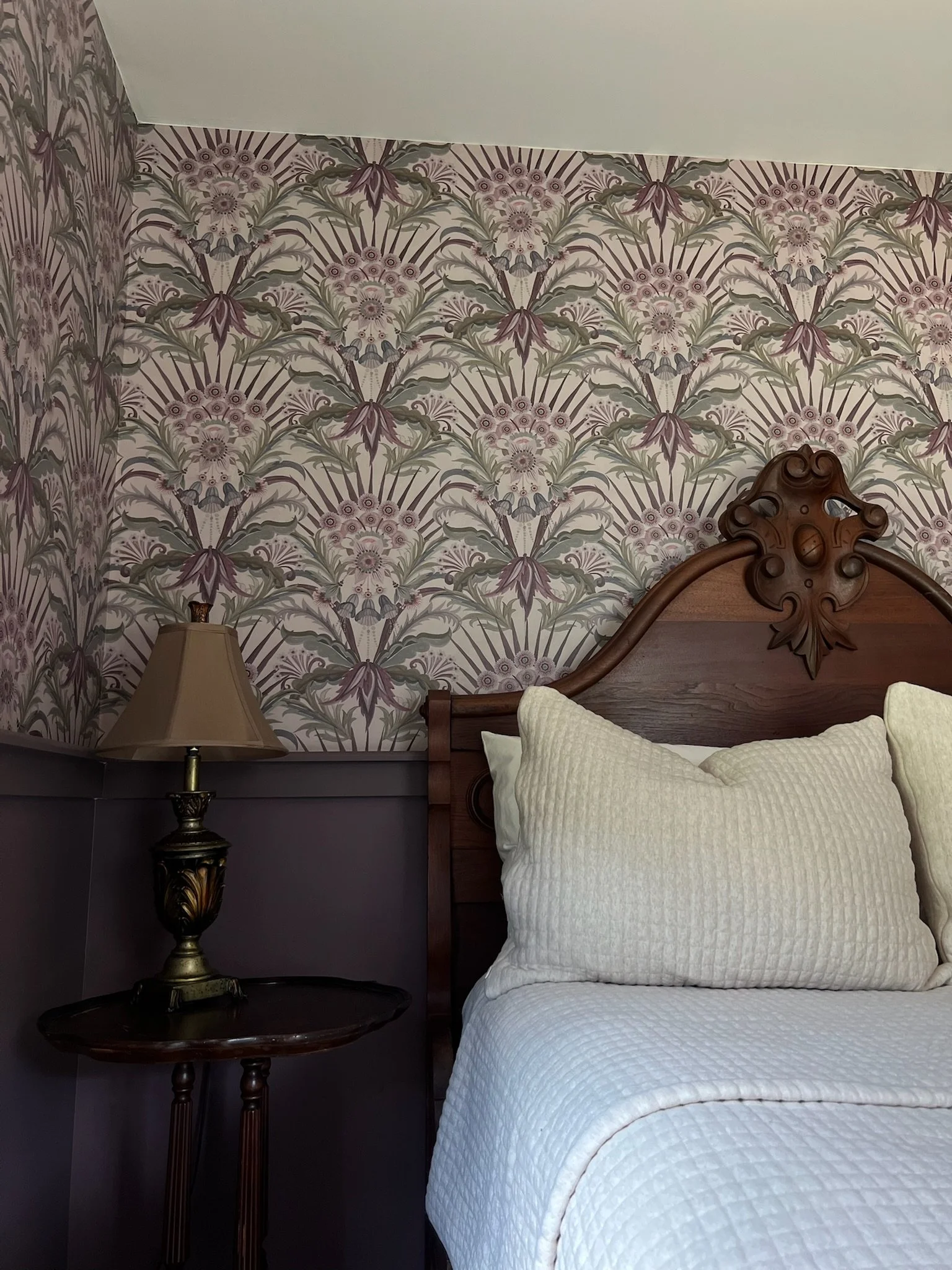 A bedroom corner featuring a wooden headboard, a bedside table with a decorative lamp, and a wall with floral-patterned wallpaper.