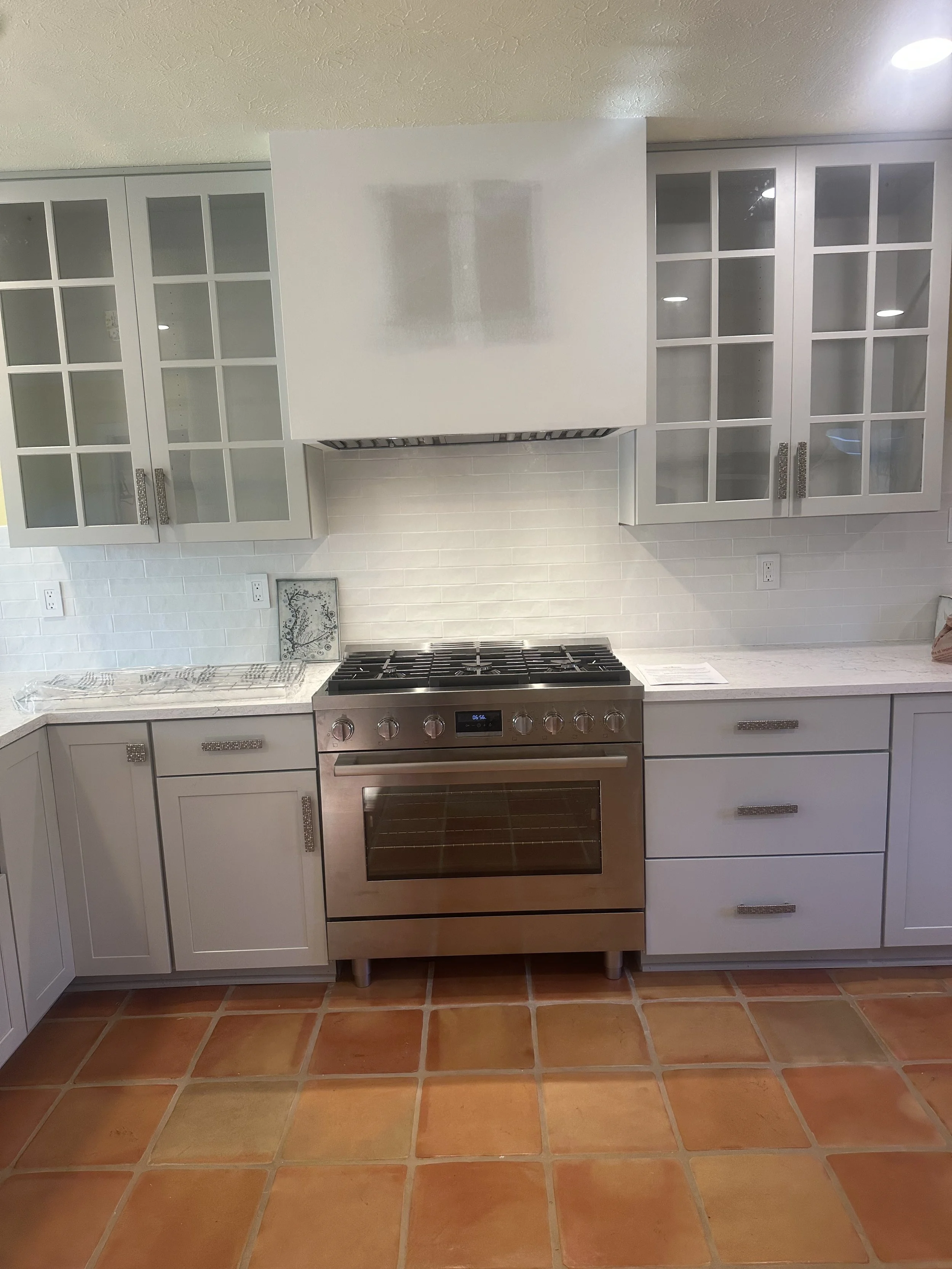 A modern kitchen with white cabinets, glass-front upper cabinets, a stainless steel stove, and orange-tiled flooring.