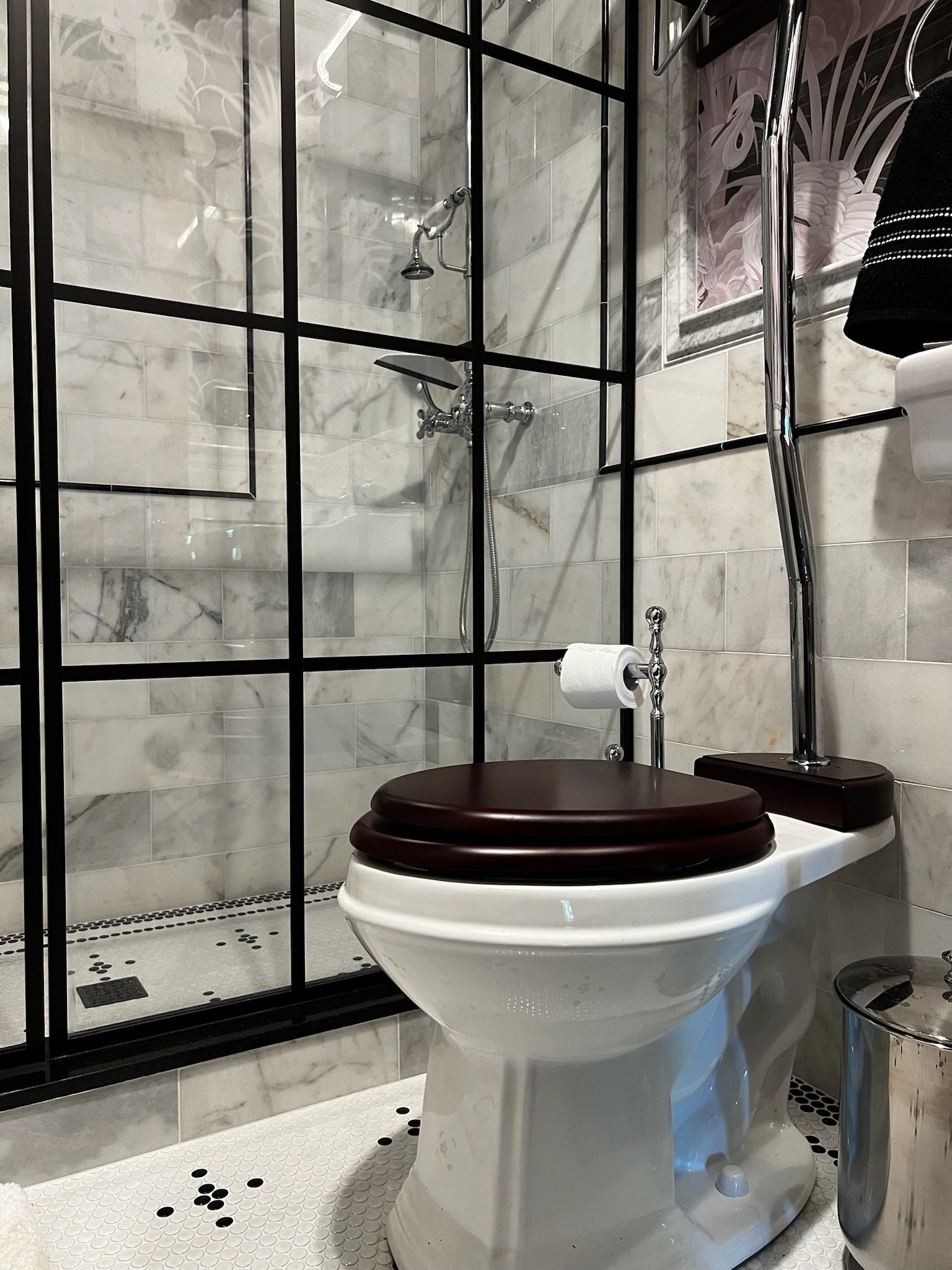 A bathroom with a glass-enclosed shower area, marble tiles, a white toilet with a dark wooden seat, a toilet paper roll, and a tiled bathroom floor with white and black mosaic tiles.