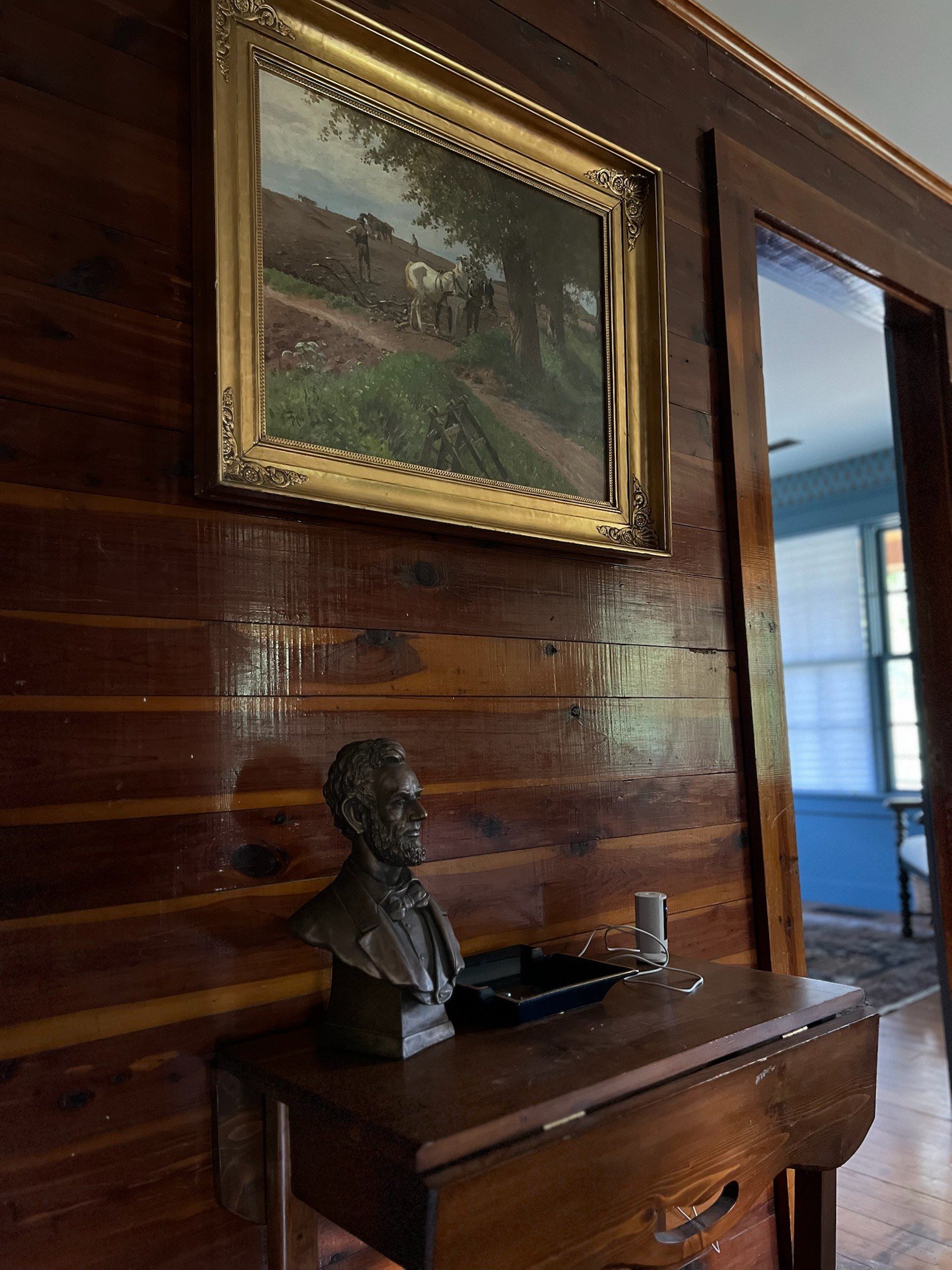 A wooden wall with a framed painting of a landscape with horses and men, a bust sculpture of a man with a beard, a wireless speaker, and a black device on a wooden table.