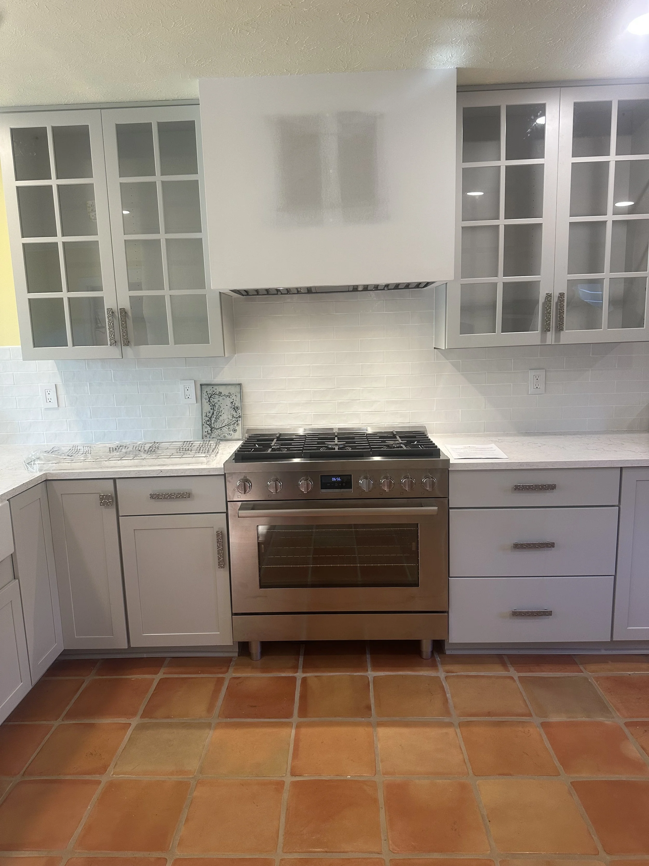 A modern kitchen with white cabinets, glass panel doors, a stainless steel oven and stove, a white tiled backsplash, and terracotta tile flooring.