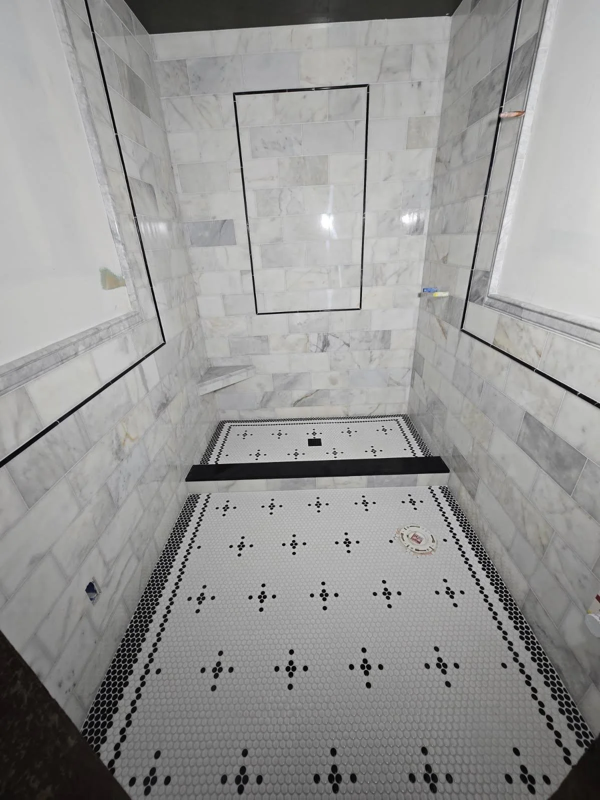 Empty shower space with white marble wall tiles, black and white mosaic floor tiles, and a black rectangular border outline on the wall.