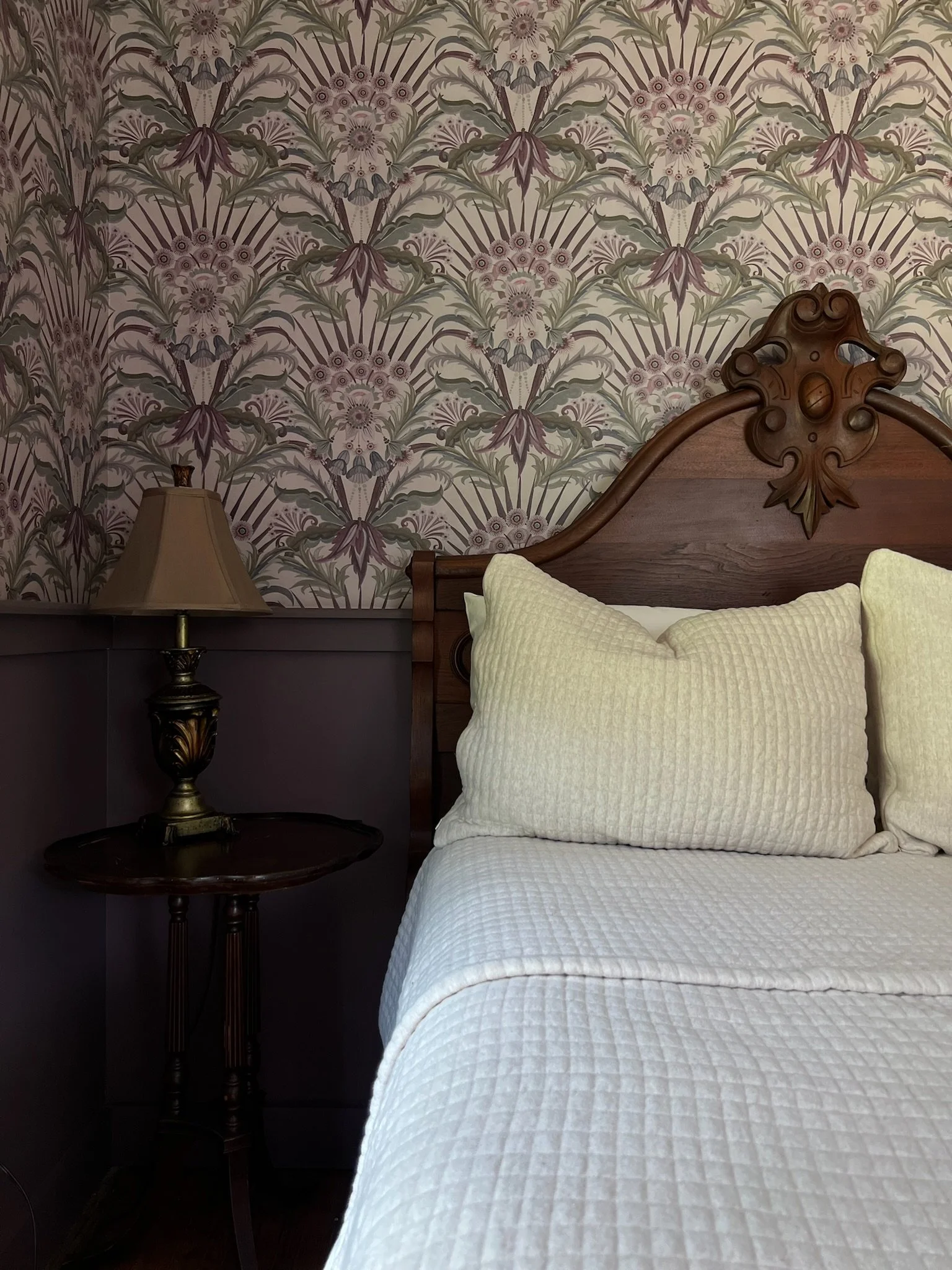 A bedroom corner with a wooden headboard, a cream-colored quilted bedspread, matching pillows, a dark wood bedside table, and a decorative table lamp. The background features floral wallpaper in pastel shades.