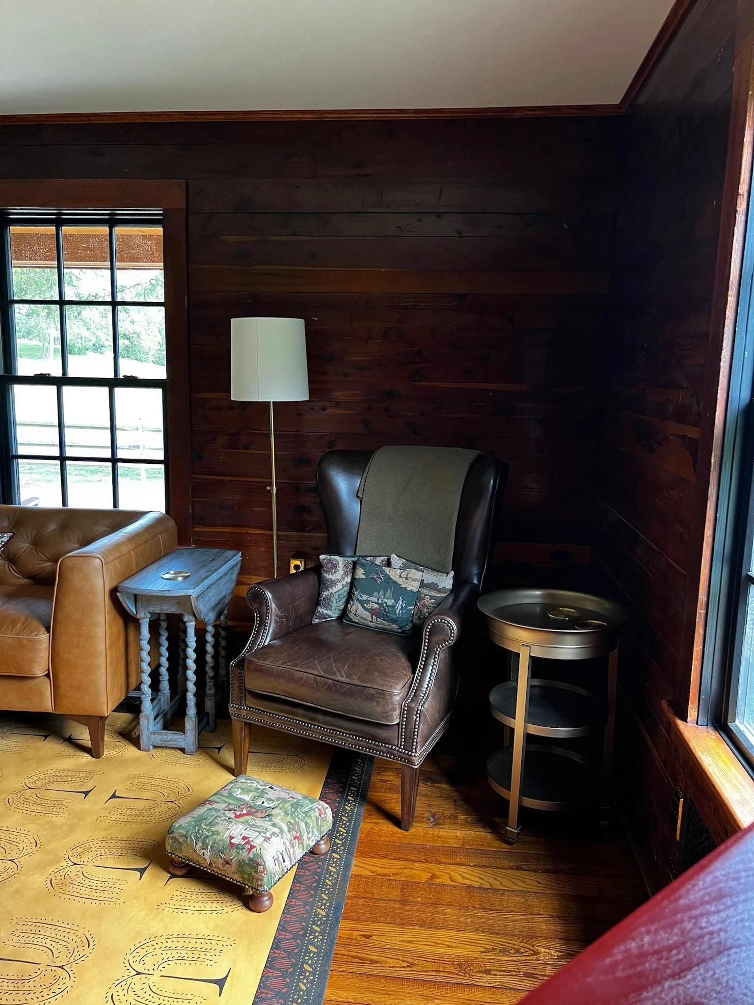 A living room corner with a leather armchair, a small side table, a floor lamp, a window with black grid, a yellow carpet with patterns, and a brown ottoman with a fabric cover. The room has dark wooden walls and hardwood floors.
