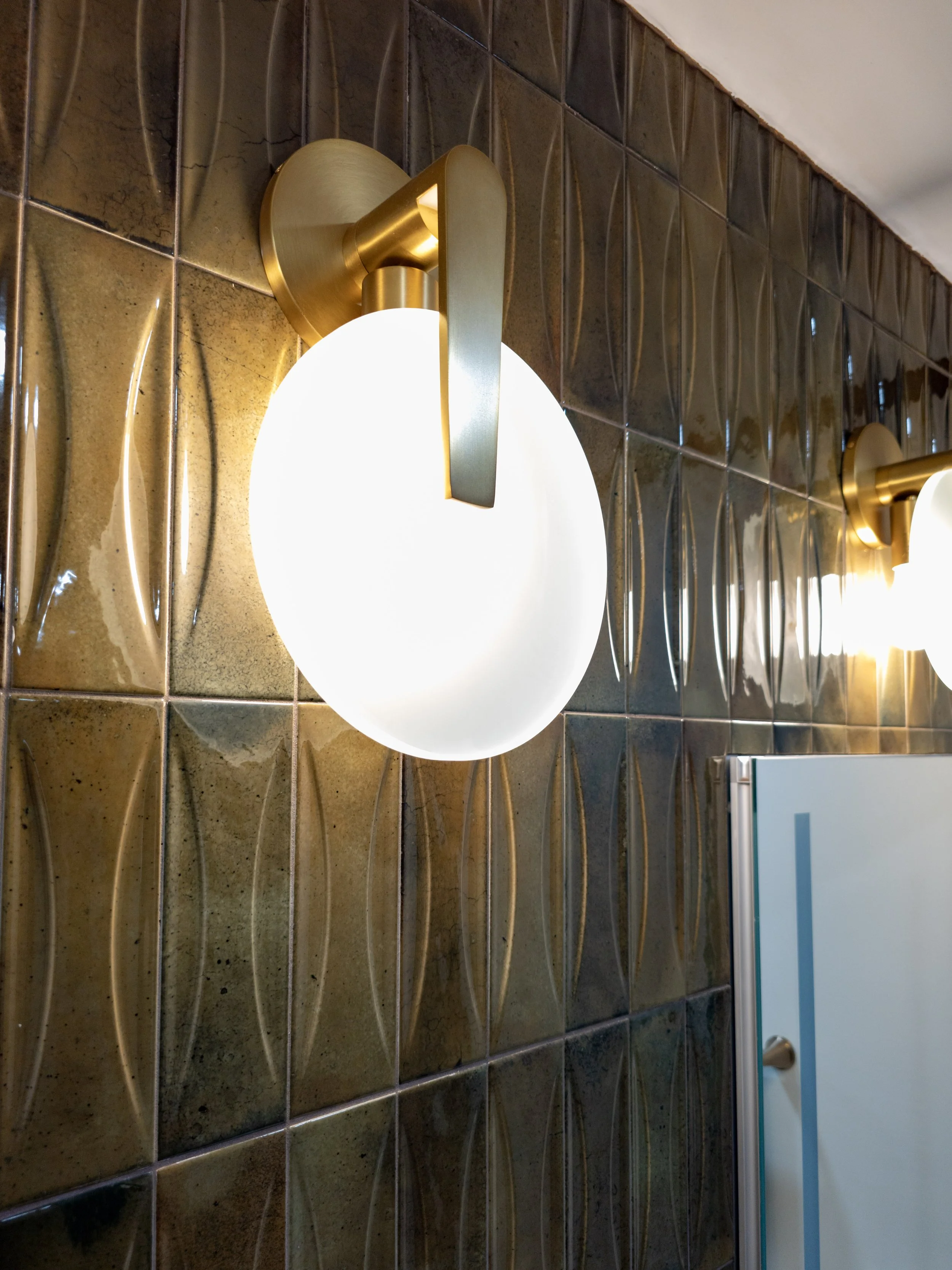 A contemporary wall-mounted light fixture with a round, white frosted glass shade and a metallic gold base, mounted on a decorative brown tiled wall.