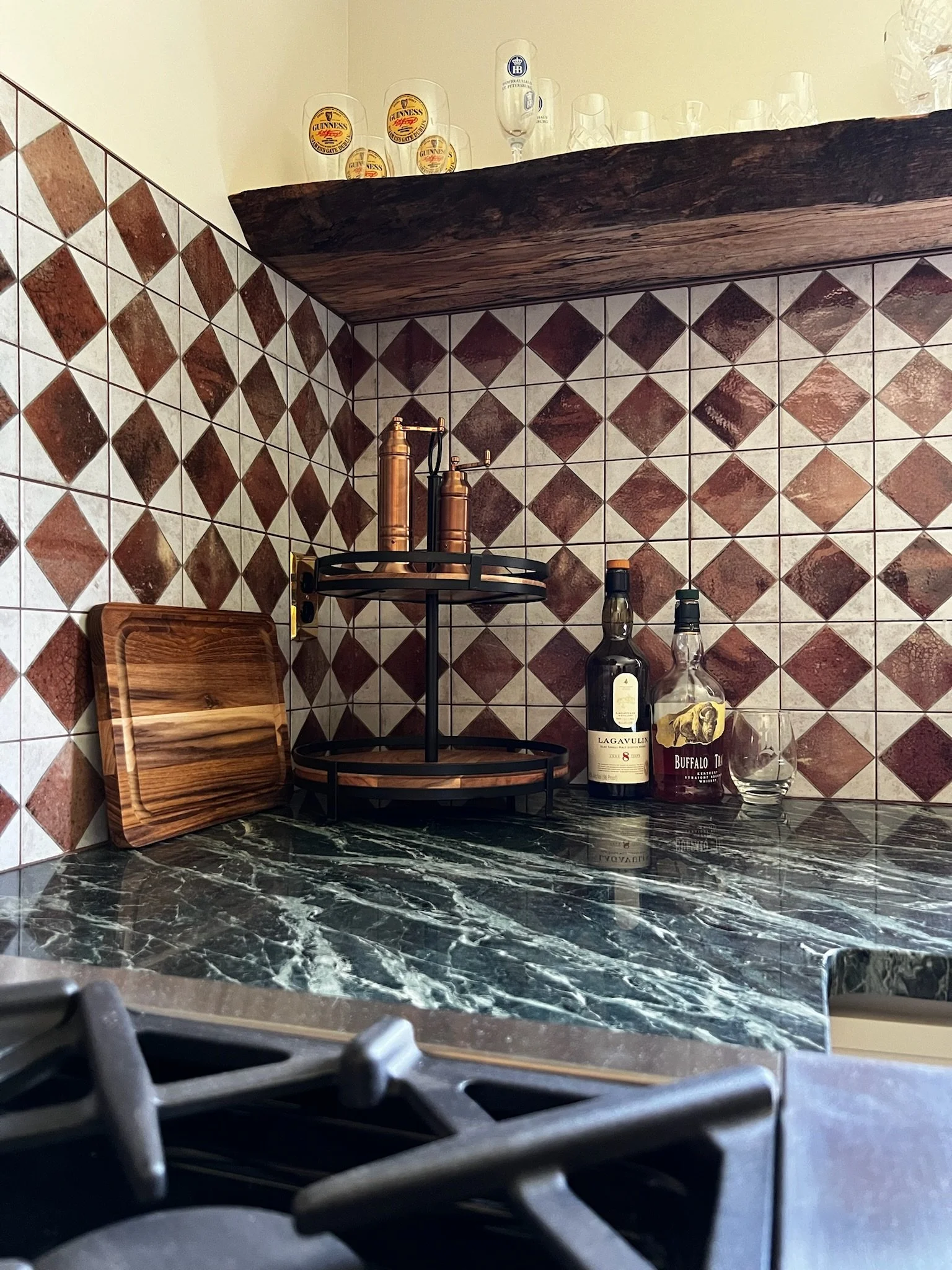 Kitchen corner with tiled wall, dark green marble countertop, wooden cutting board, and bottles of alcohol. Glasses are on a wooden shelf above.