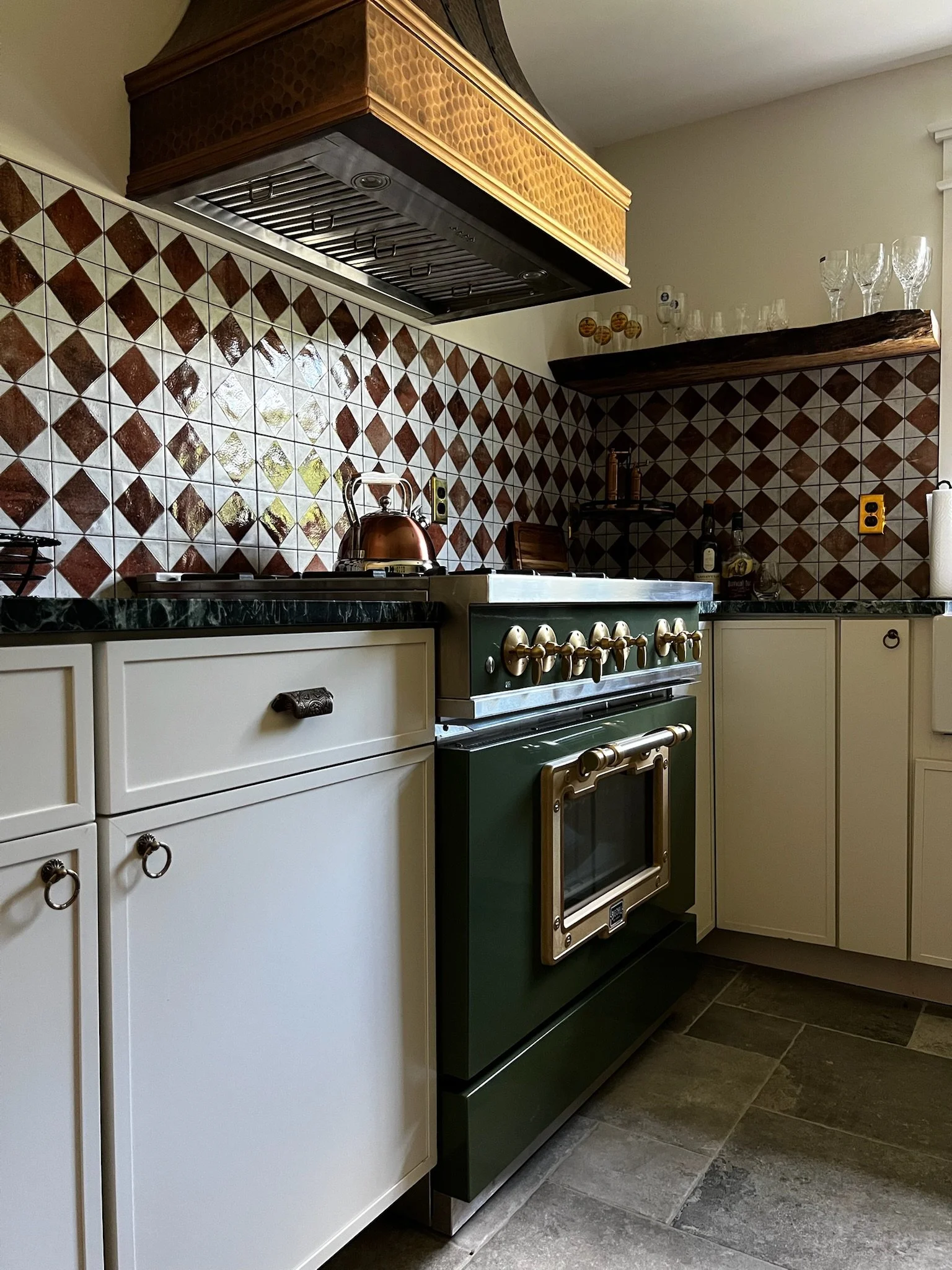 Kitchen with a vintage green stove, white cabinets, a copper kettle, a dark green countertop, and a tiled backsplash with a diamond pattern in brown and white. Shelves hold glassware and bottles.