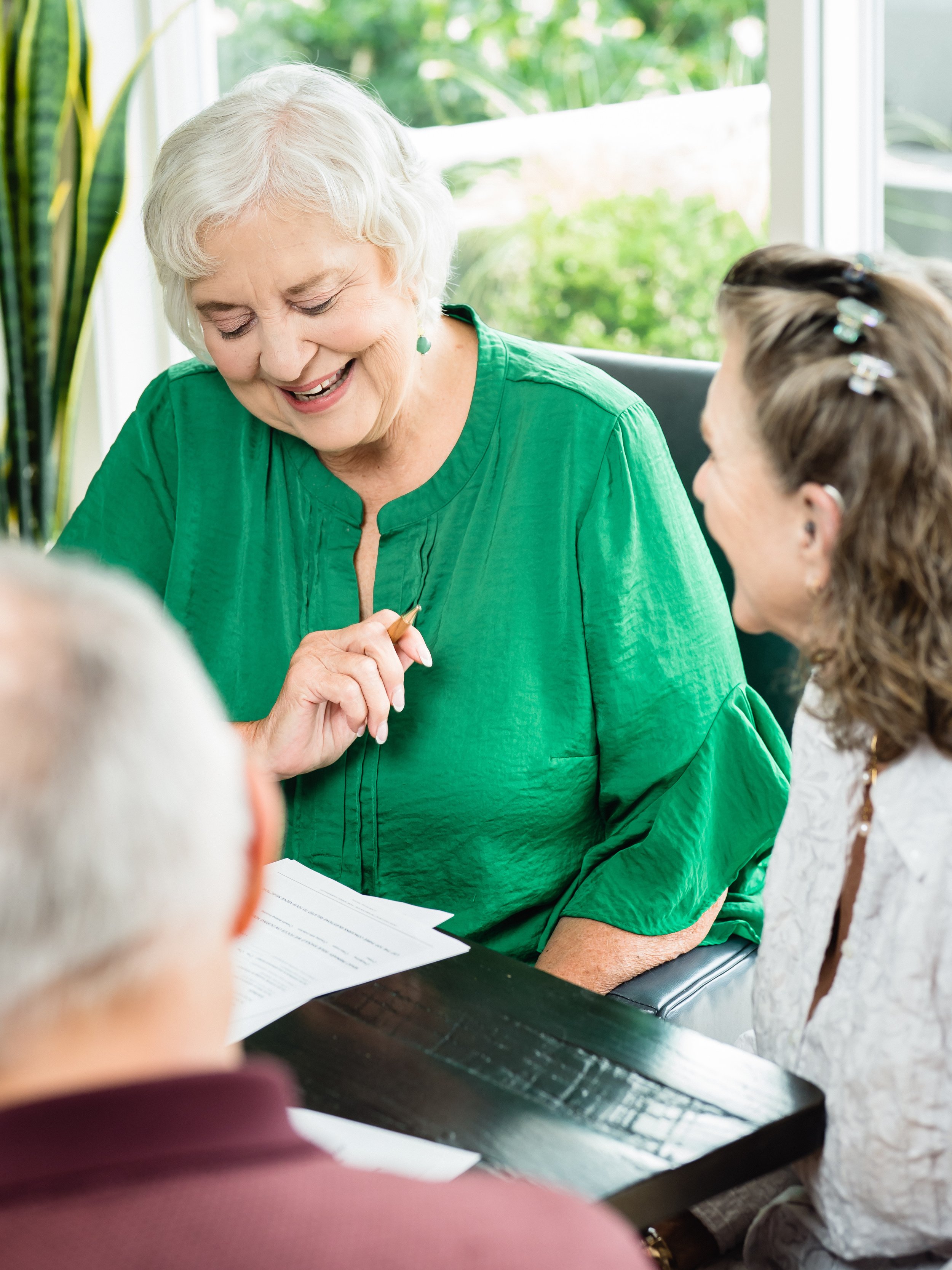 A smiling elderly woman in a green blouse sitting at a table with three other people, reading a document.