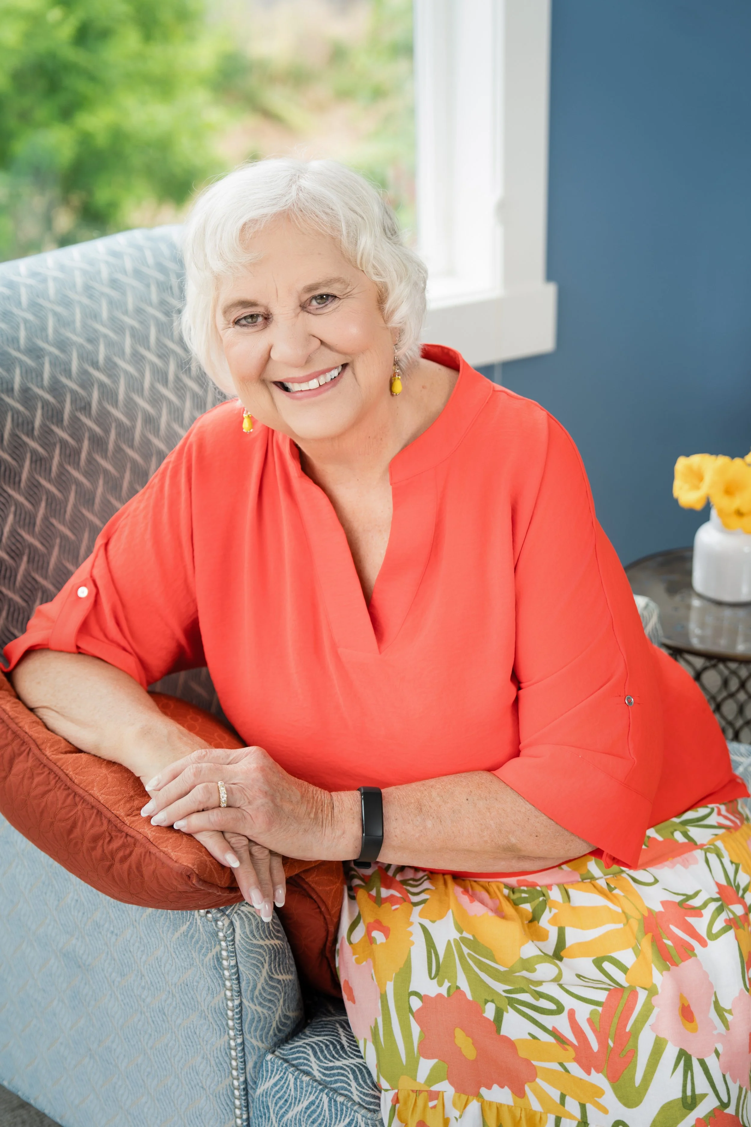 Smiling elderly woman with short white hair, wearing an orange top and floral skirt, sitting on a couch with a window and greenery in the background.