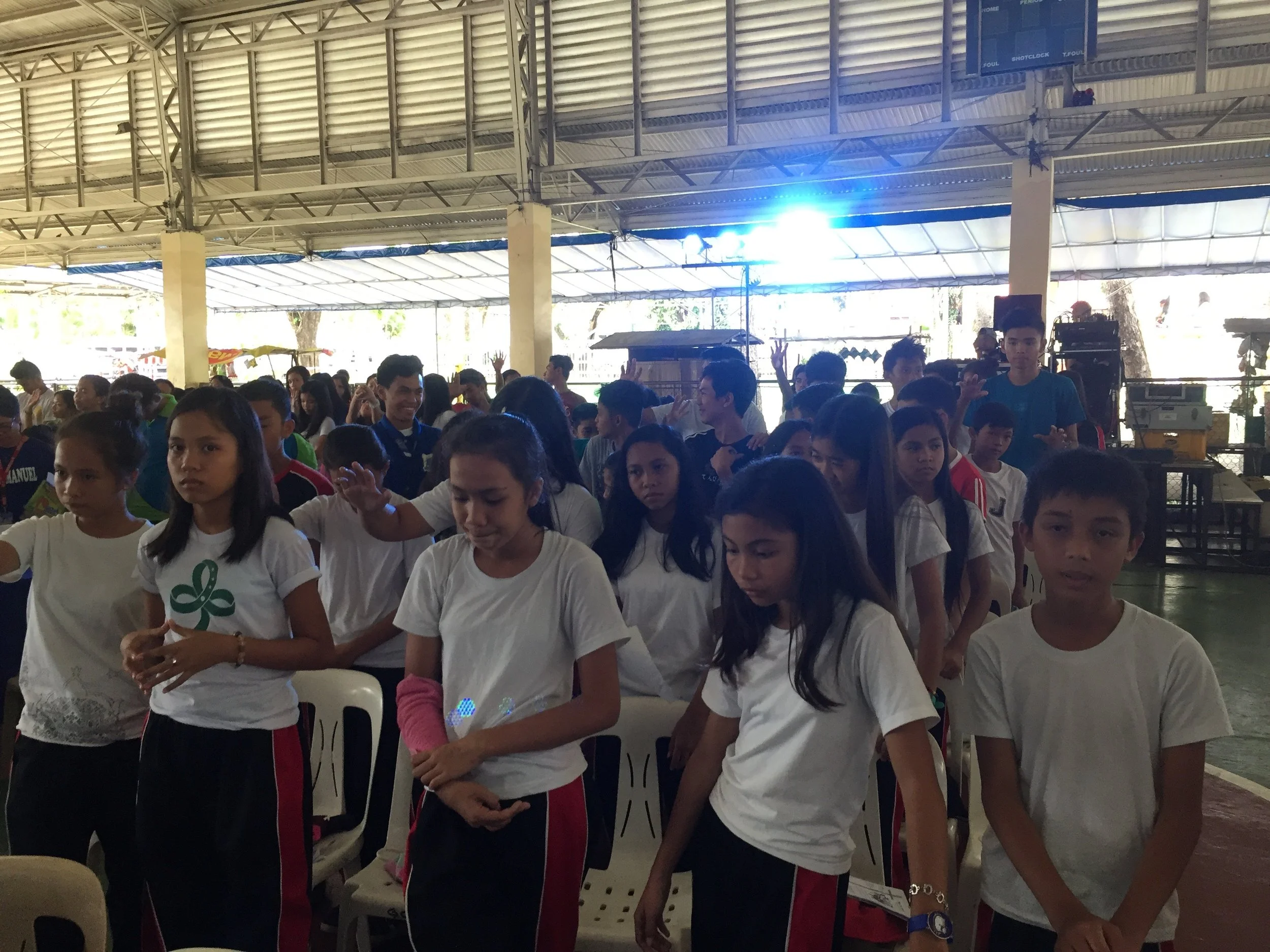 Group of children and teenagers gathered indoors, some with hands raised, participating in an event or activity.