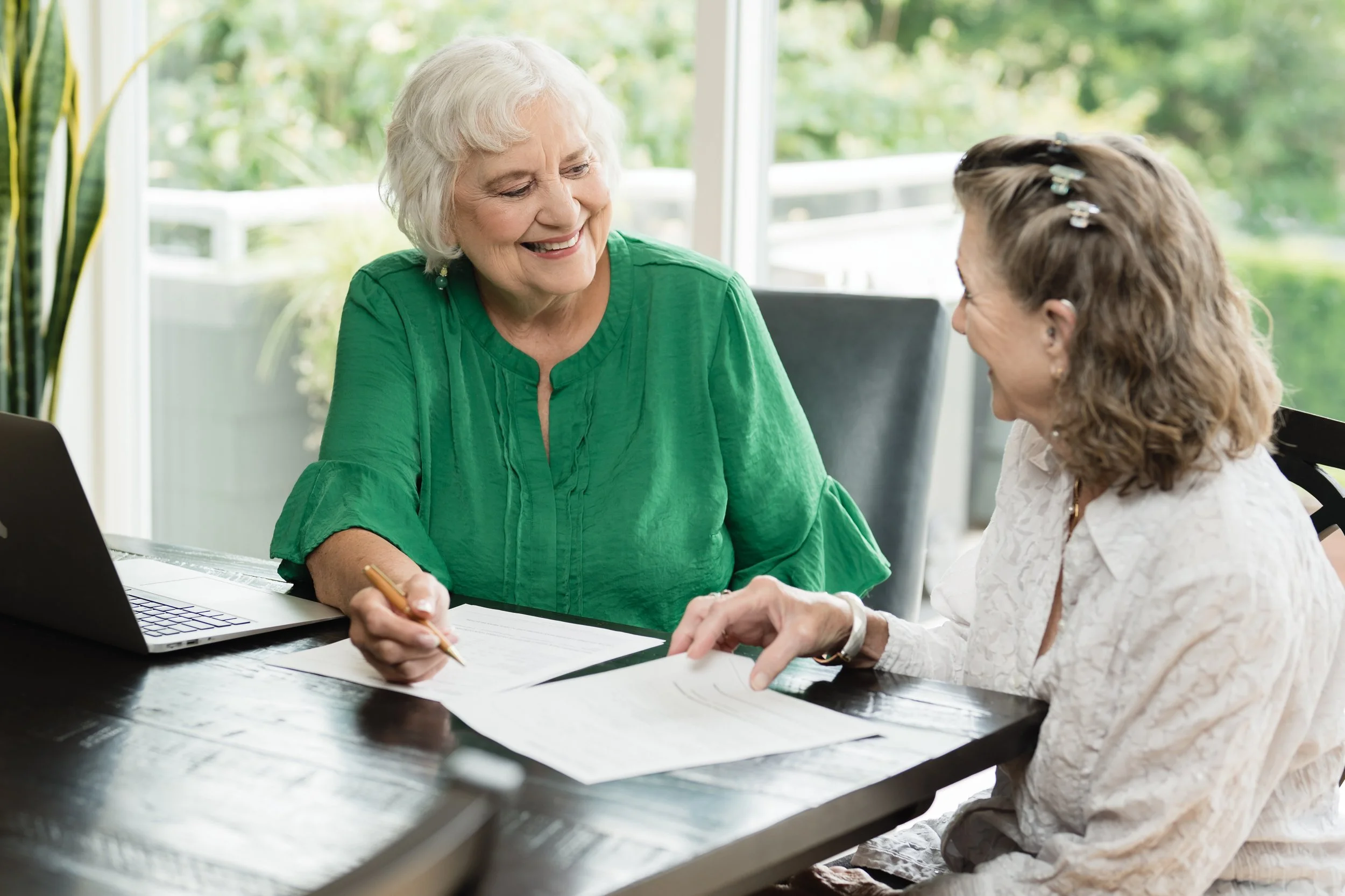 Two women sitting at a table, smiling and engaging in a conversation, with papers and a laptop in front of them, in a bright room with large windows and greenery outside.