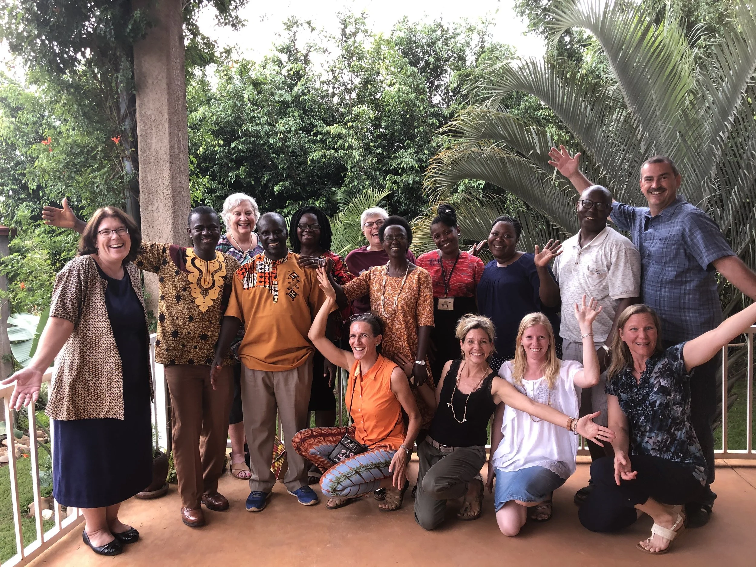Group of diverse adults smiling and posing on a balcony with lush green trees and plants in the background.
