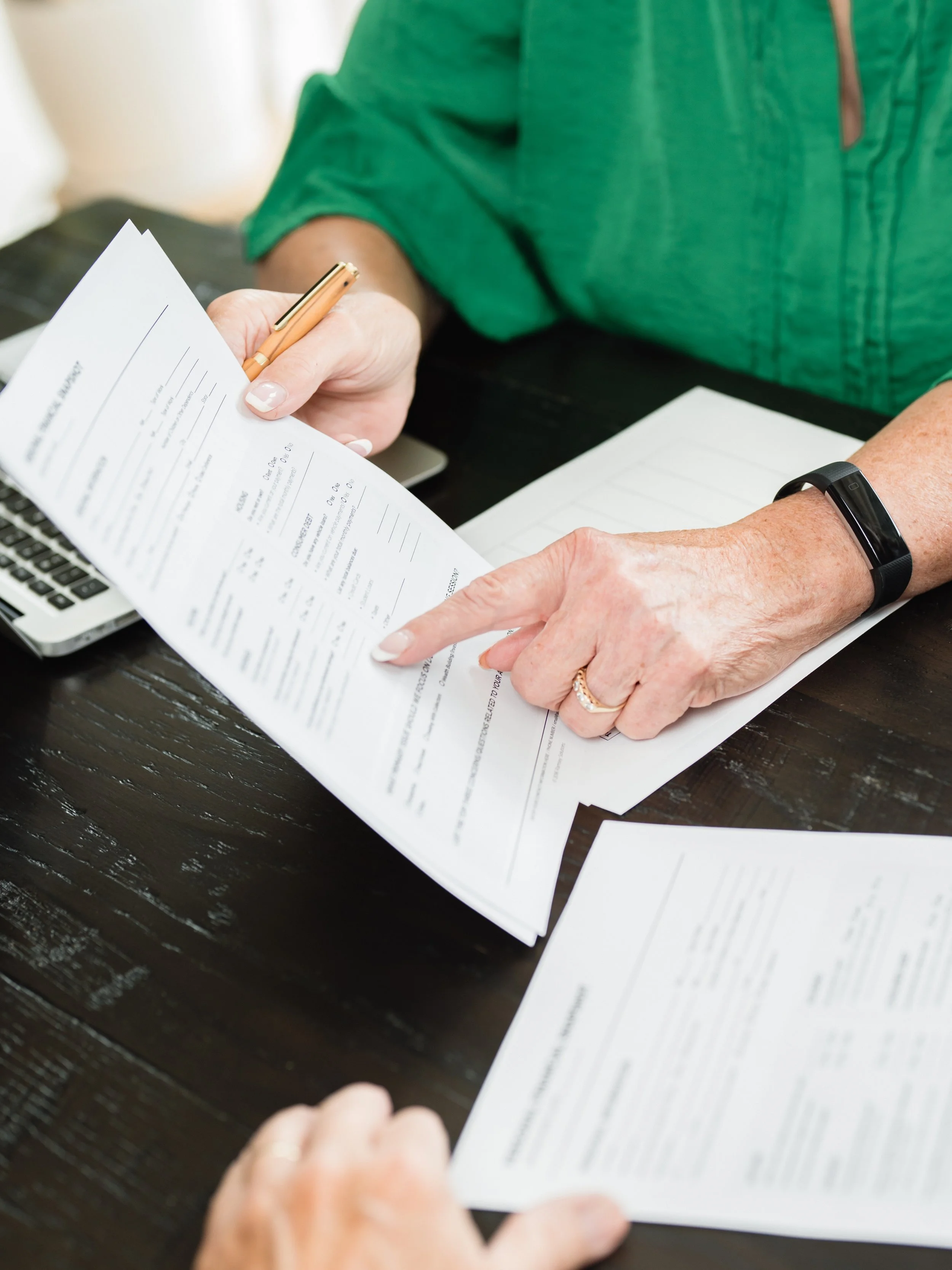 Close-up of a person reviewing and pointing at a printed document, with a pen in hand, on a dark wooden table.
