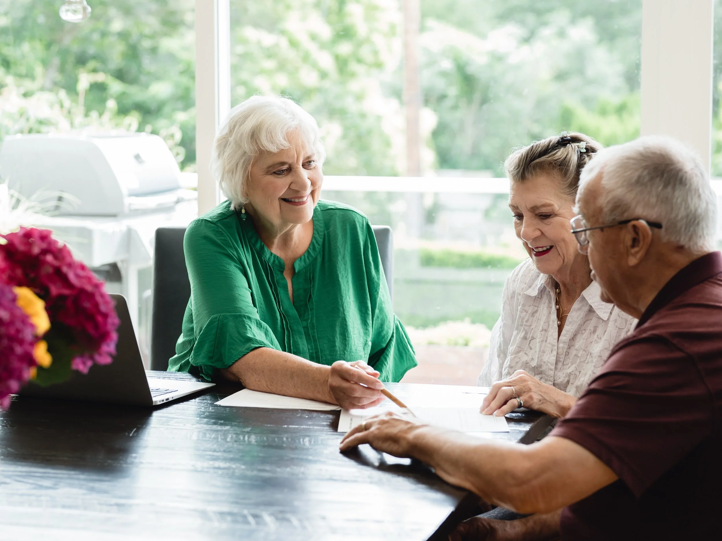 Three senior adults, two women and one man, engaged in a discussion at a table, with the woman in a green shirt smiling and pointing at a document, while the other two listen attentively.