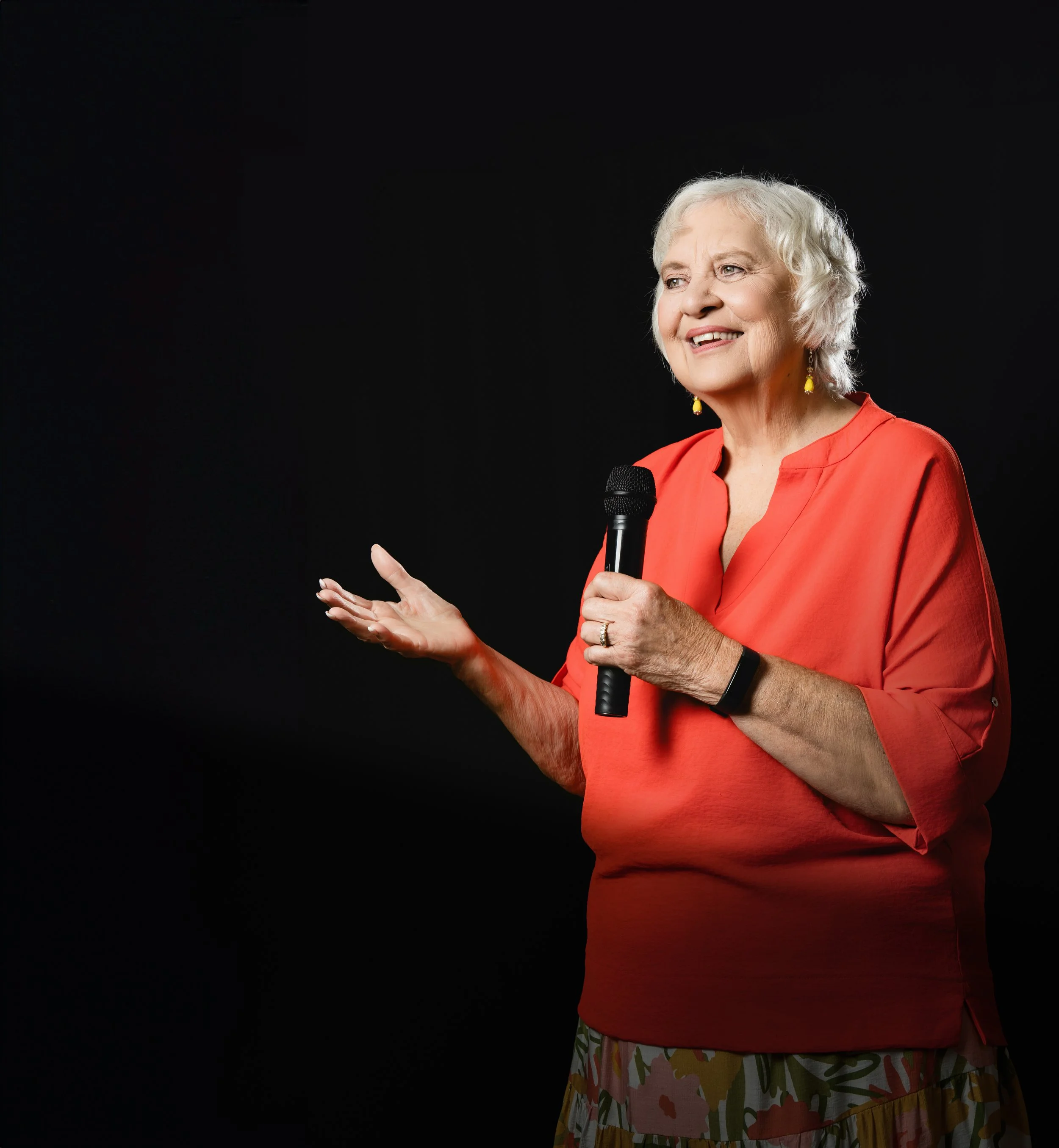Older woman with white hair holding a microphone, gesturing with her left hand, wearing a red blouse and earrings, standing against a black background.