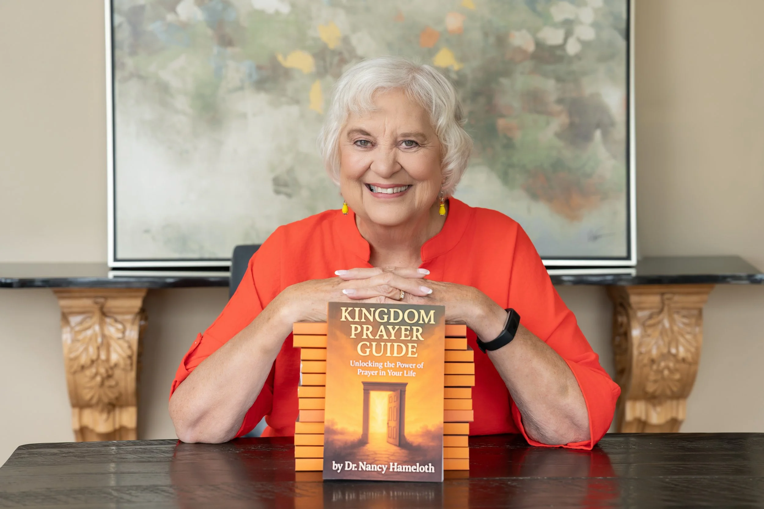 An elderly woman with white hair wearing a red blouse and yellow earrings sitting at a table with a book titled "Kingdom Prayer Guide" by Dr. Nancy Hameloth in front of her. She is smiling and resting her cheecks on her clasped hands. A large TV screen and a decorative wooden shelf are in the background.