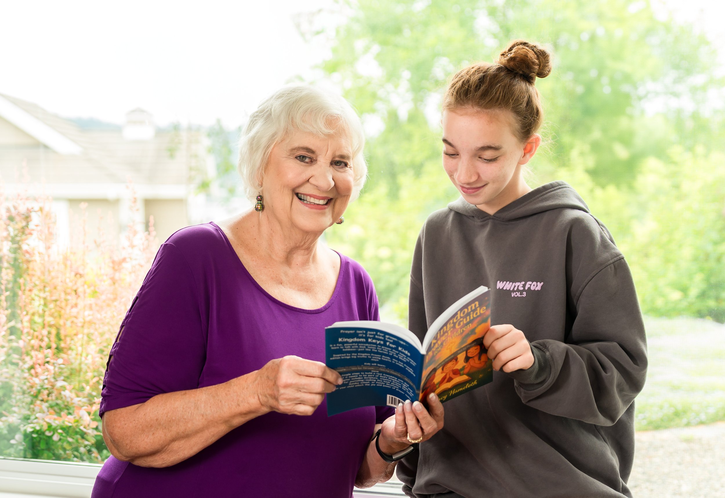 An elderly woman and a teenage girl stand by a window, smiling while reading a book together on a bright day with green trees outside.