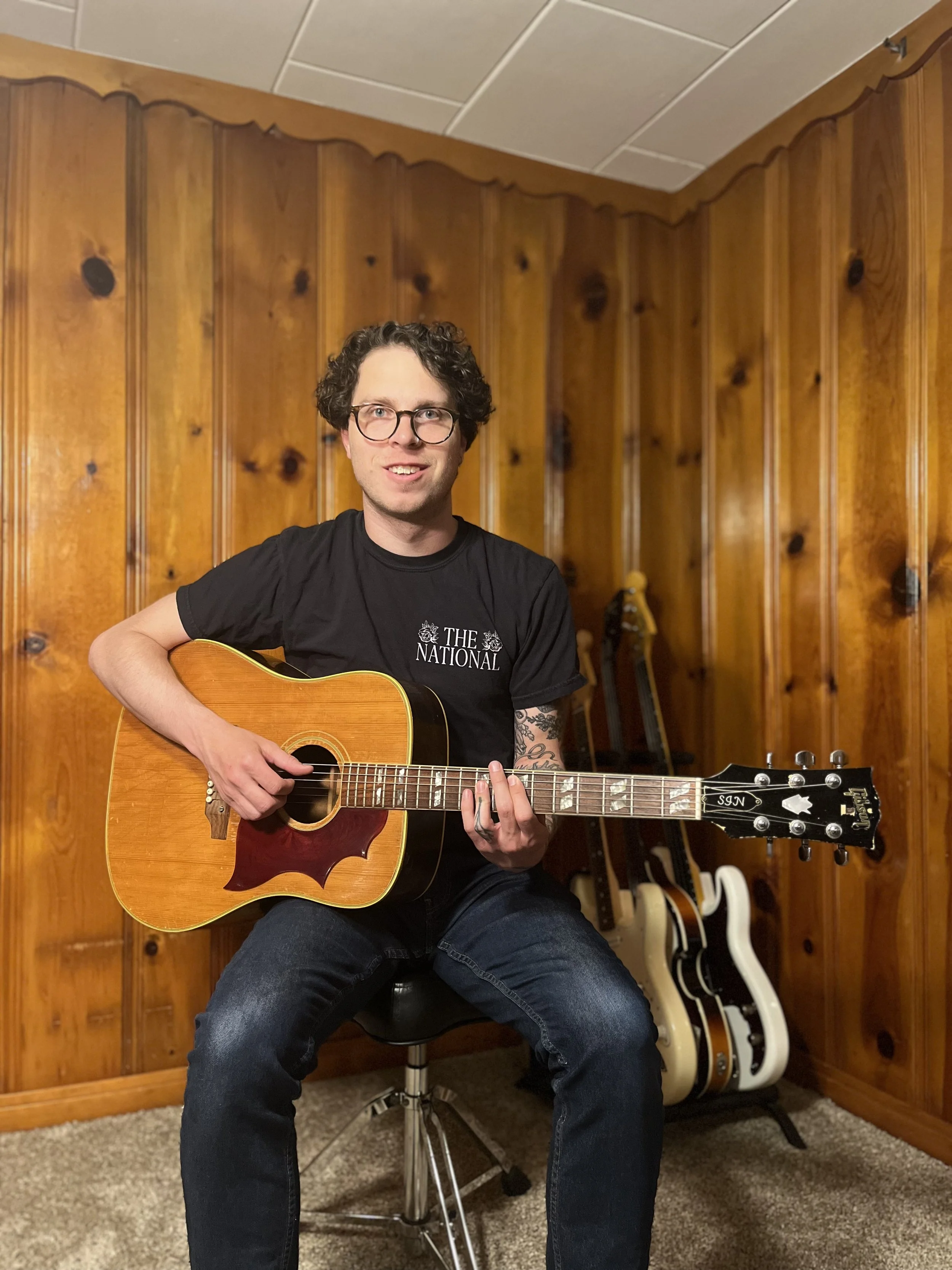 A man with curly hair and glasses sitting on a stool, taking guitar lessons in Denver.