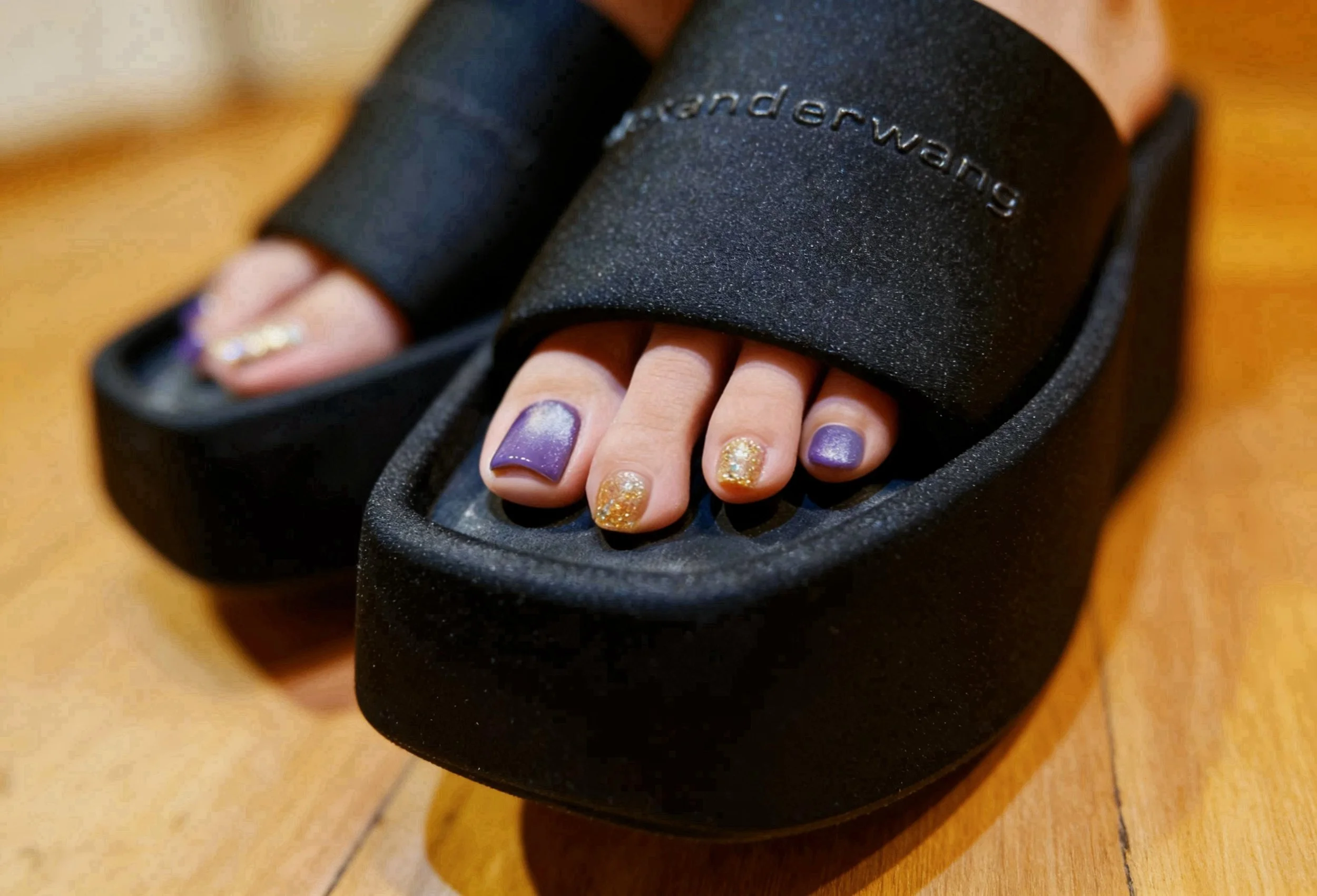Close-up of toes with purple and glittery gold nail polish inside a black foot care device labeled "underwand" on a wooden surface.