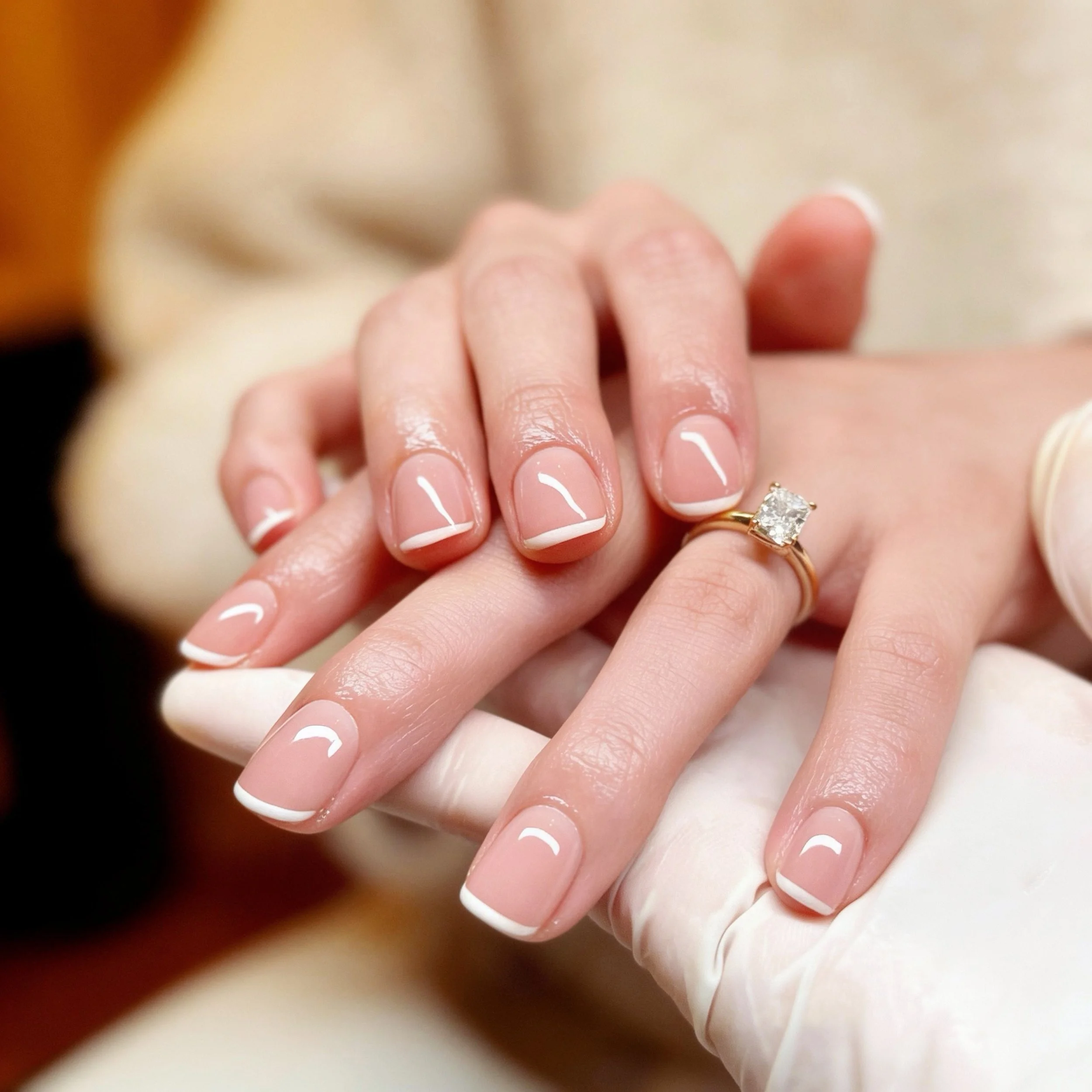 Close-up of a hand with well-manicured nails resting on another hand with a gold ring with a large diamond. The background is blurred.