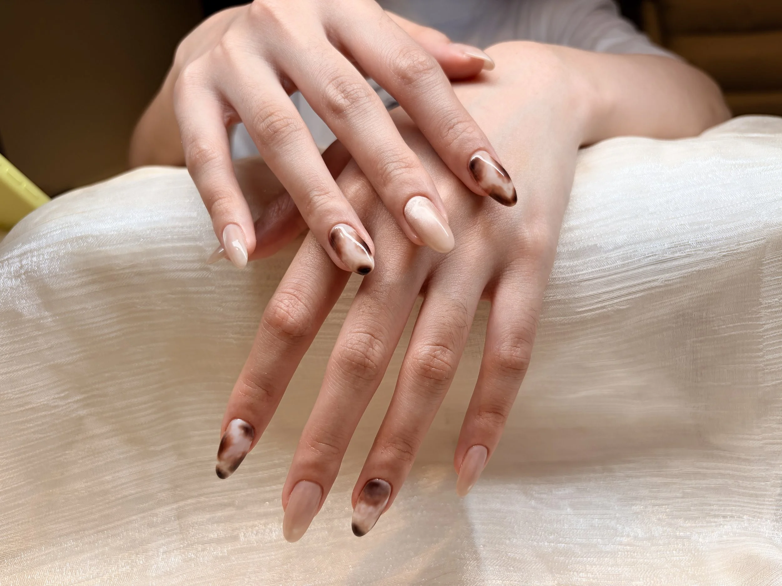 Close-up of two hands resting on a satin fabric, with one hand on top of the other. The nails are styled with a swirling marble pattern in neutral tones.