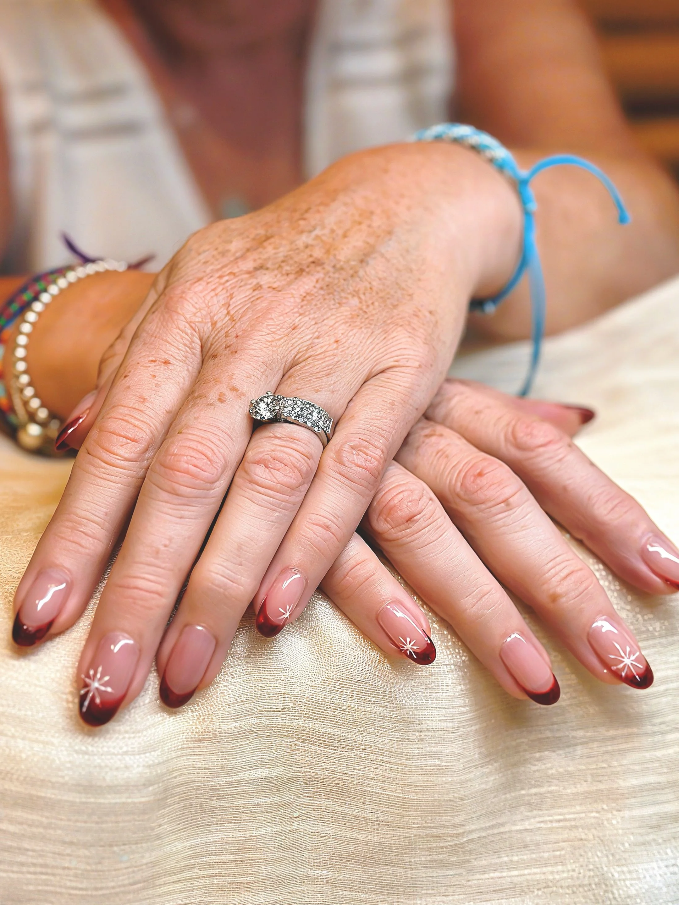 Close-up of a woman's hand with manicured nails and a diamond ring, resting on a satin surface. The nails are painted with a red-gradient design.