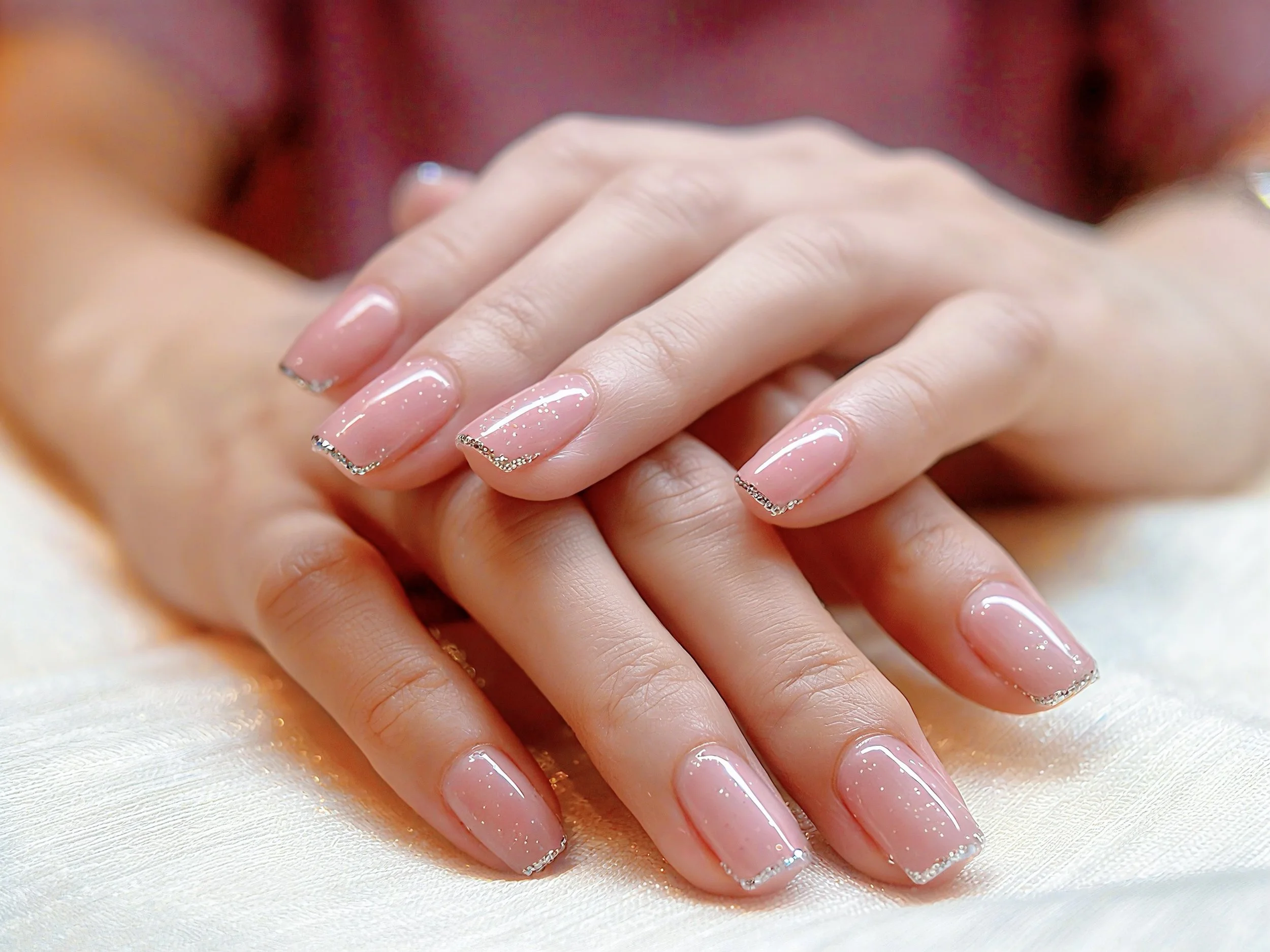 Close-up of elegantly manicured nails with a clear shiny polish and delicate silver rhinestone accents along the tips, resting on a soft, light-colored fabric.