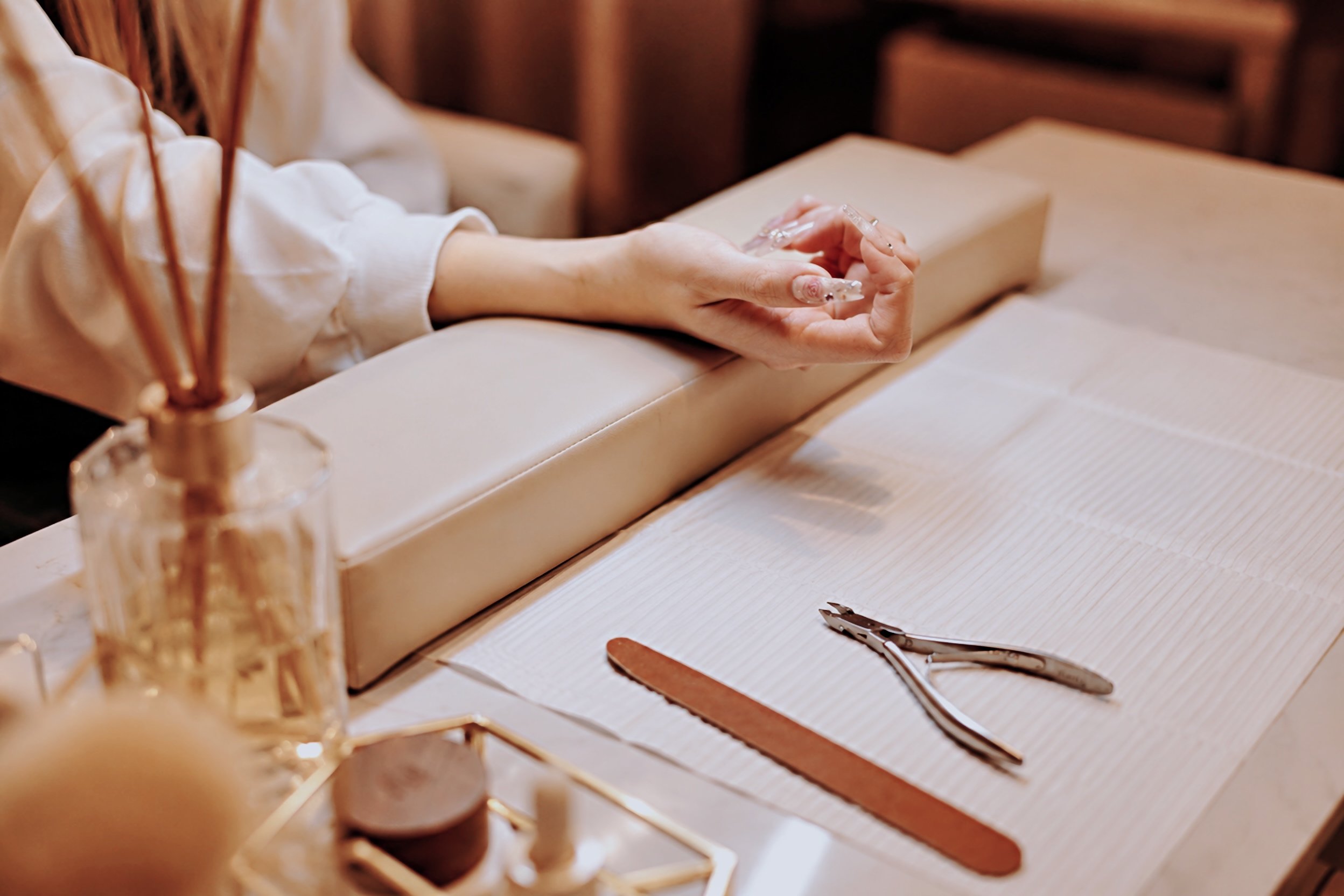 Close-up of a person's hands holding a glass nail file, with manicure tools on a table. In the background, there is a beige-colored leather surface and blurred objects.