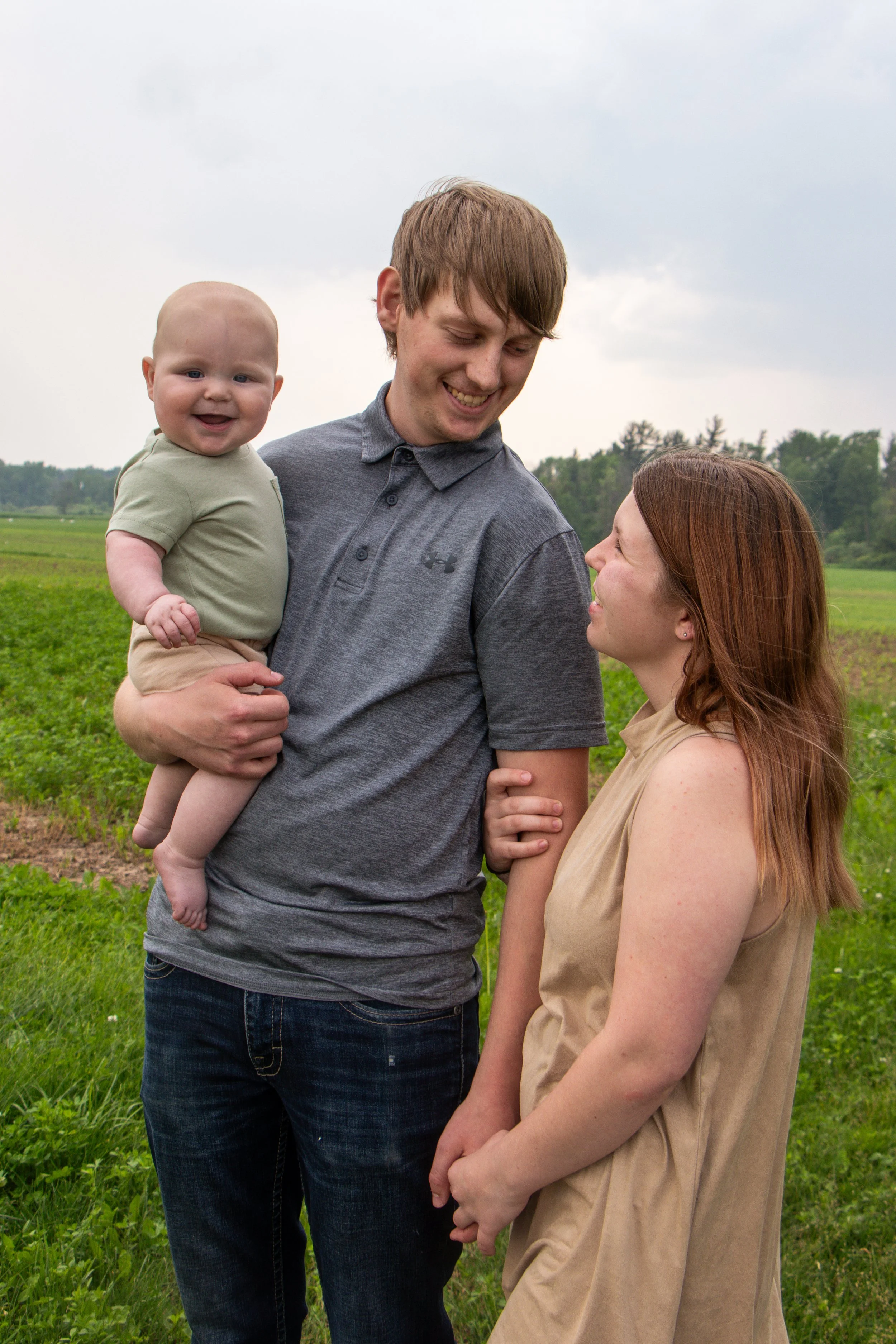 A young man holding a smiling baby boy in a field, looking down and smiling at a woman with long red hair who is looking up at him.