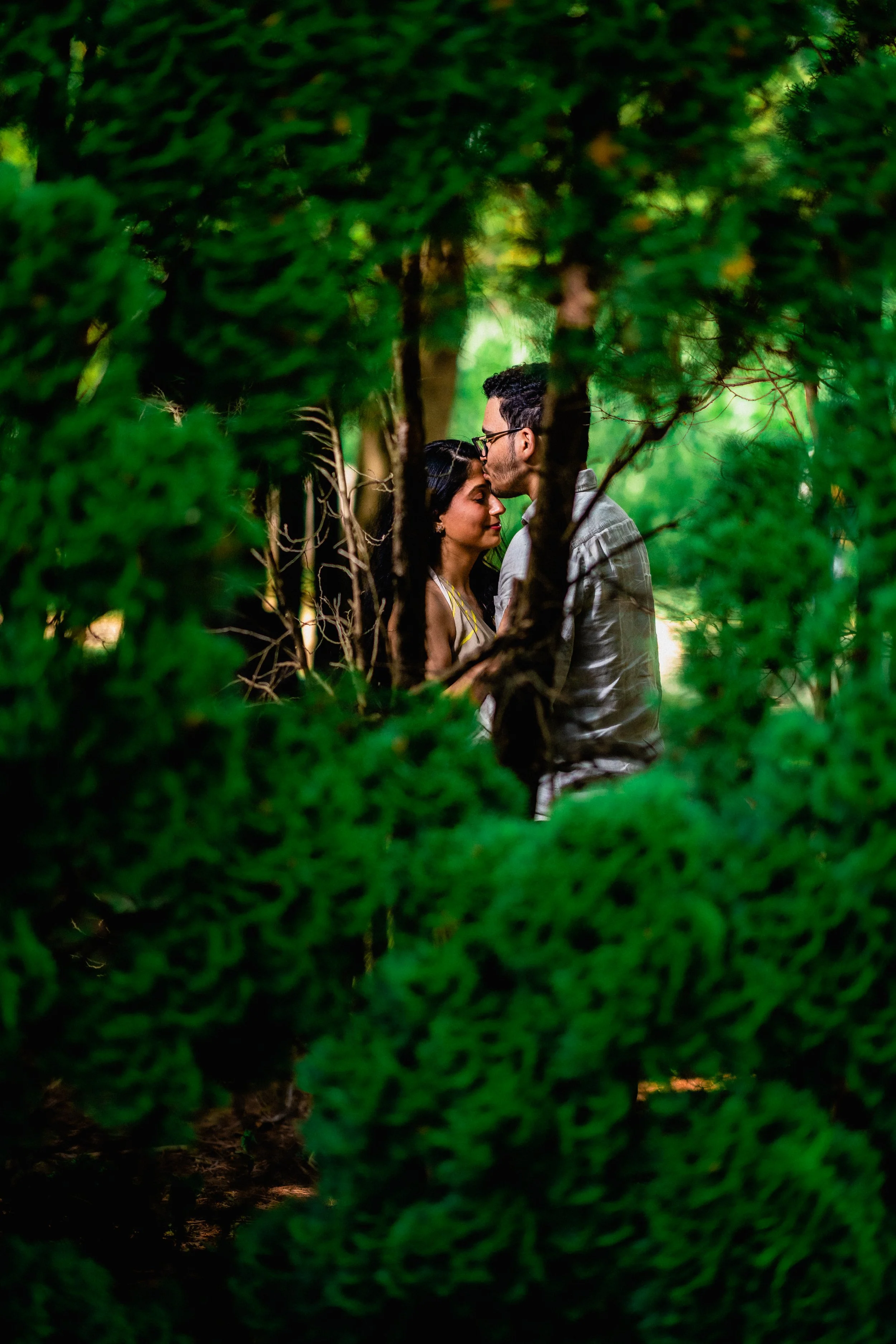 NJ Wedding Photography of A man and a woman share a tender moment in a green, wooded area, surrounded by lush branches and foliage.