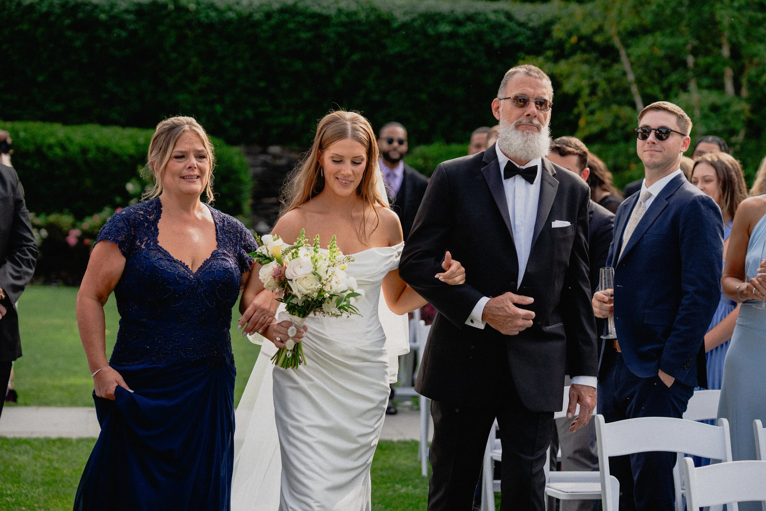 Documentary wedding photography of a bride walking down the outdoor aisle with family, surrounded by guests and greenery.