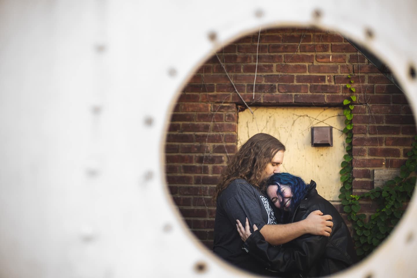 Two people embrace, seen through a circular opening, standing against a brick wall with green vines, expressing affection and happiness.