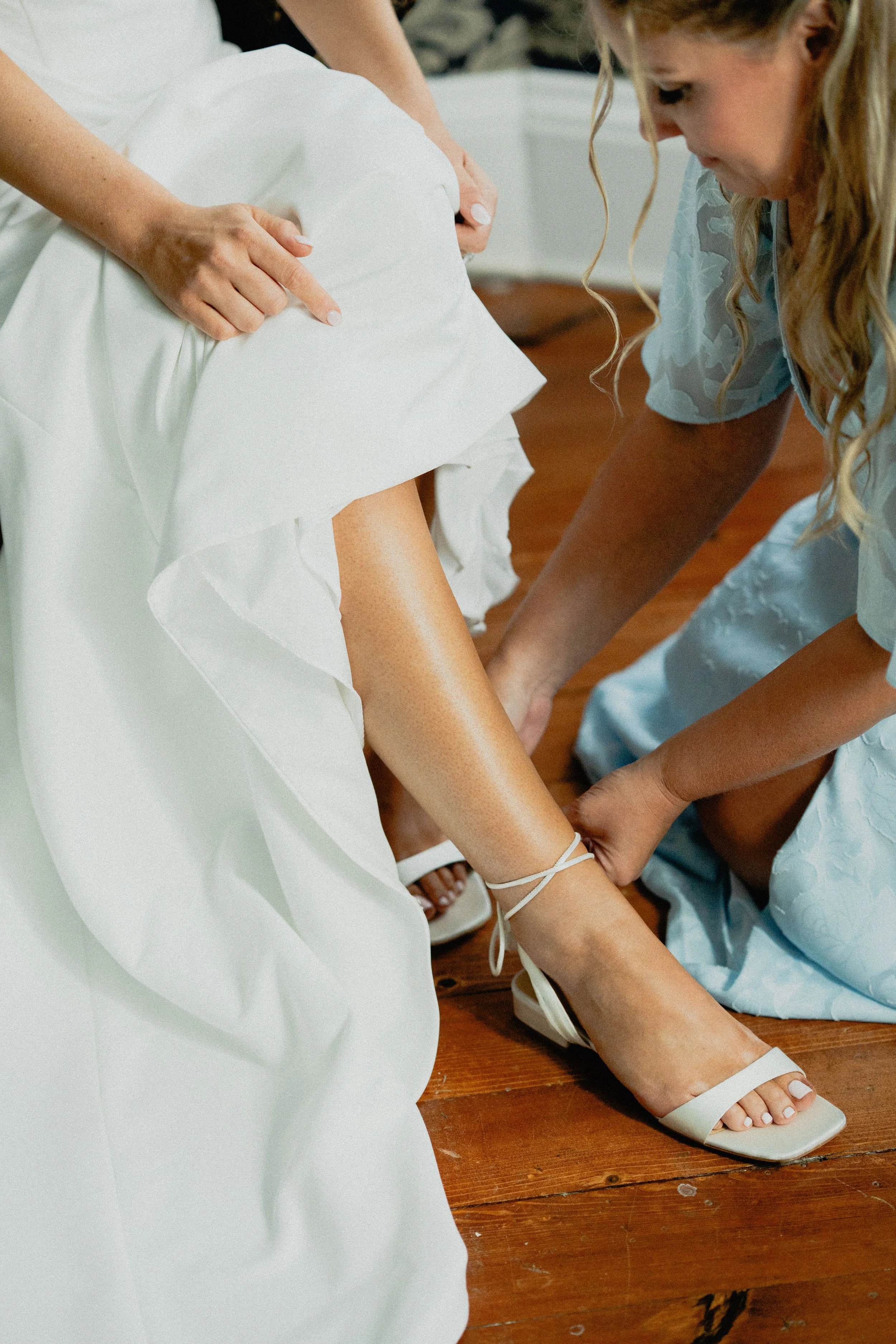 Documentary wedding photography of a bridesmaid fastening the bride's white heels during wedding-day preparations.