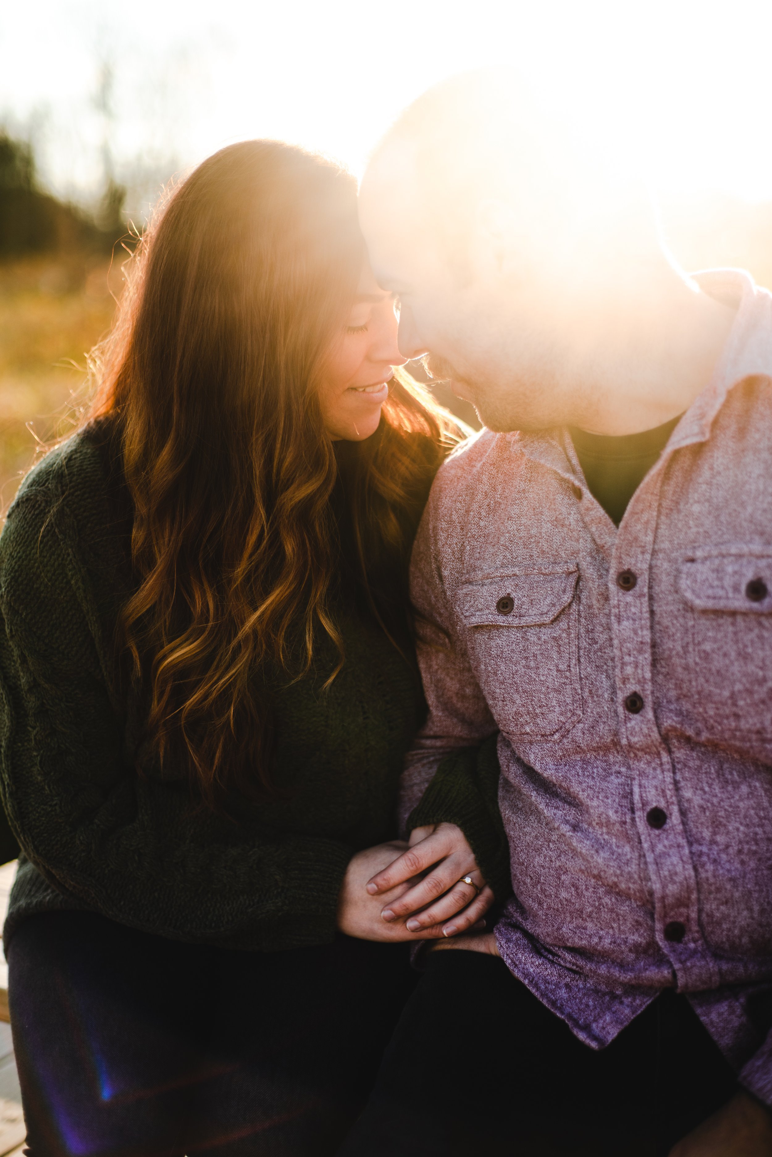 A couple with their foreheads and noses touching, outdoors during sunset, with the sunlight backlighting them.
