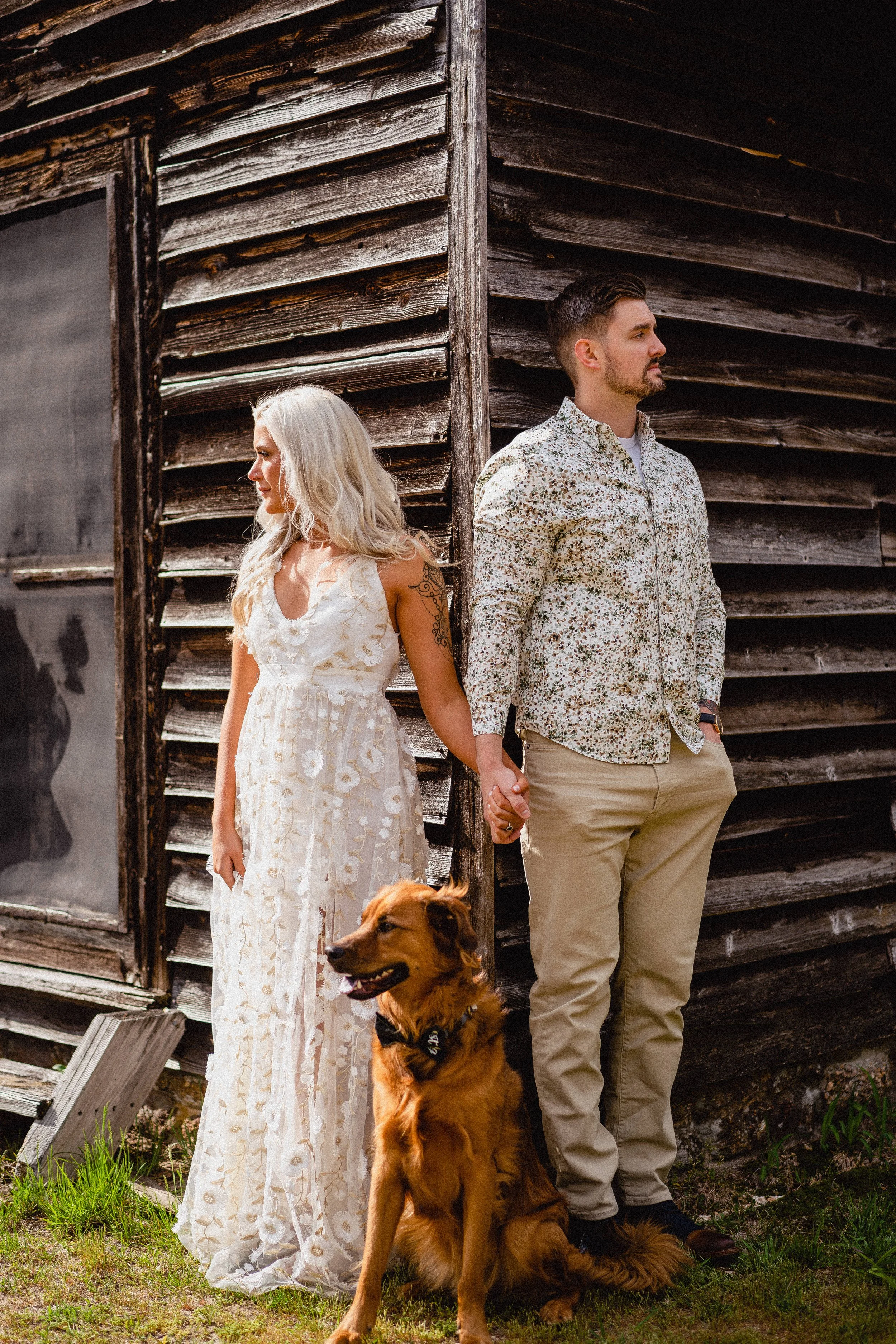 NJ Wedding Photography of A man and woman holding hands outdoors, standing back to back near a weathered wooden building, with a golden retriever sitting in front of them.
