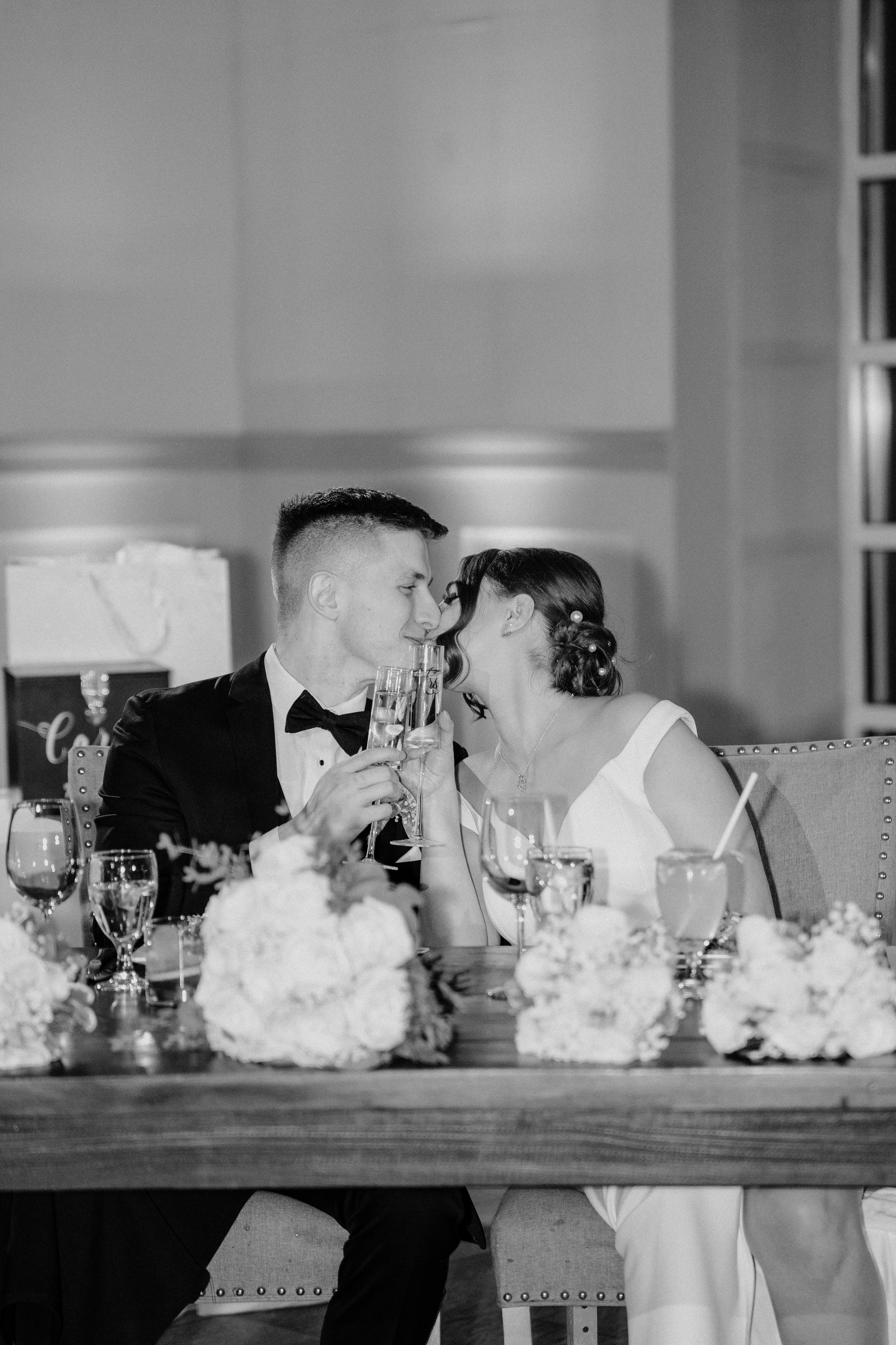 Black and white photo of a bride and groom sharing a kiss at their wedding reception, sitting at a decorated table with flowers and drinks.