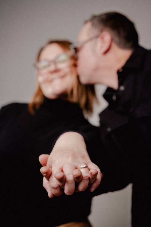 Close-up of a couple holding hands with an engagement ring, woman with red hair and glasses, man with short hair, both smiling, woman wearing black top, affectionate moment.