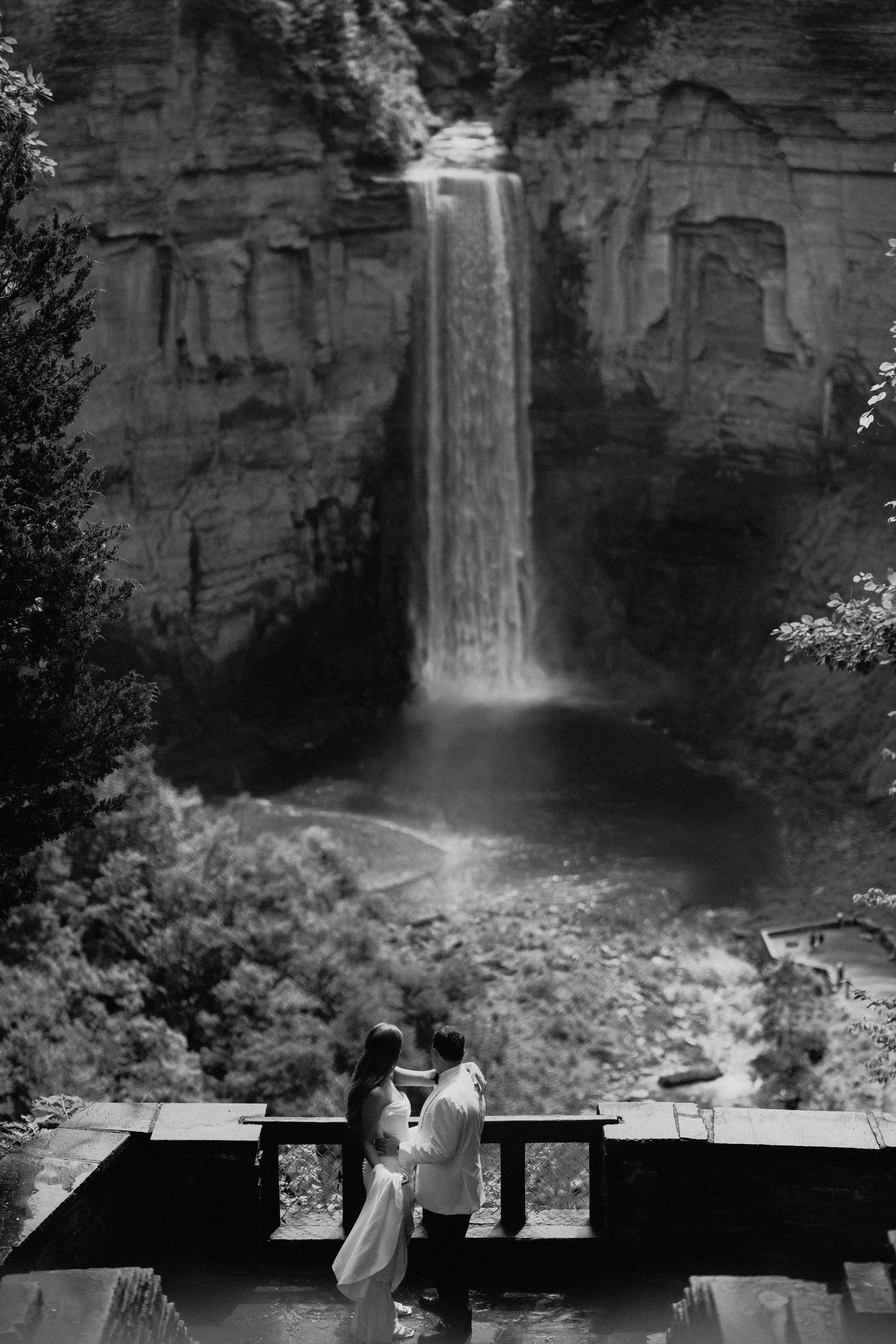 Black-and-white editorial wedding photography portrait of a bride and groom on a balcony overlooking a rocky waterfall.