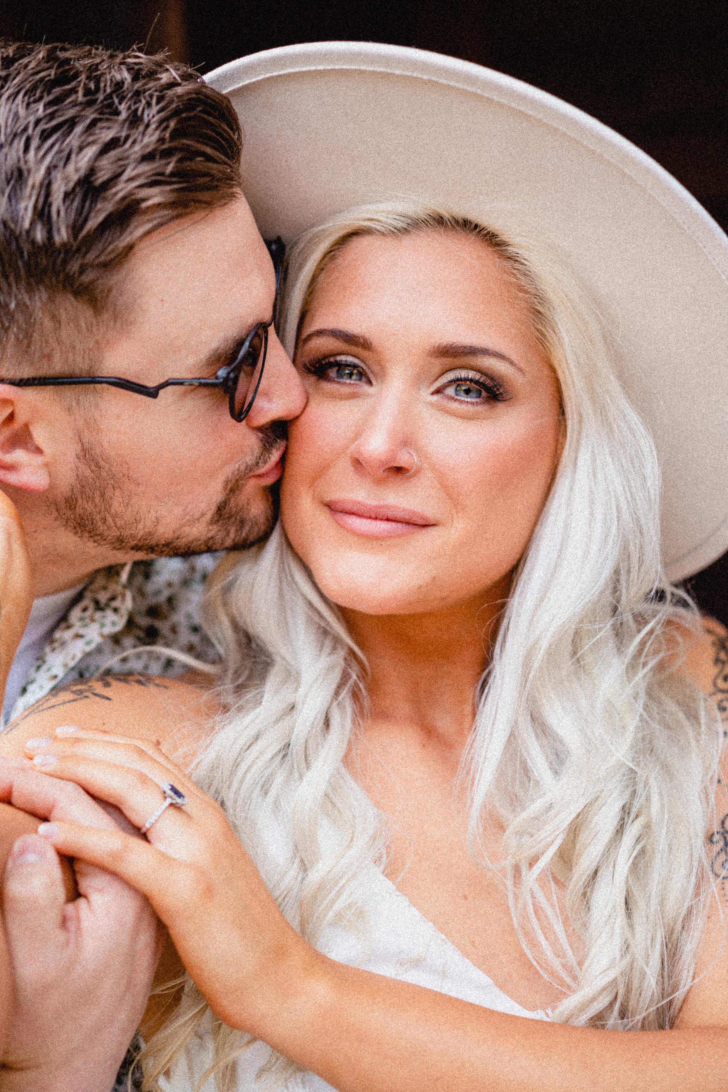 A man with glasses and a woman with long blonde hair, wearing a white hat, in an intimate close-up shot, gazing softly at the camera.