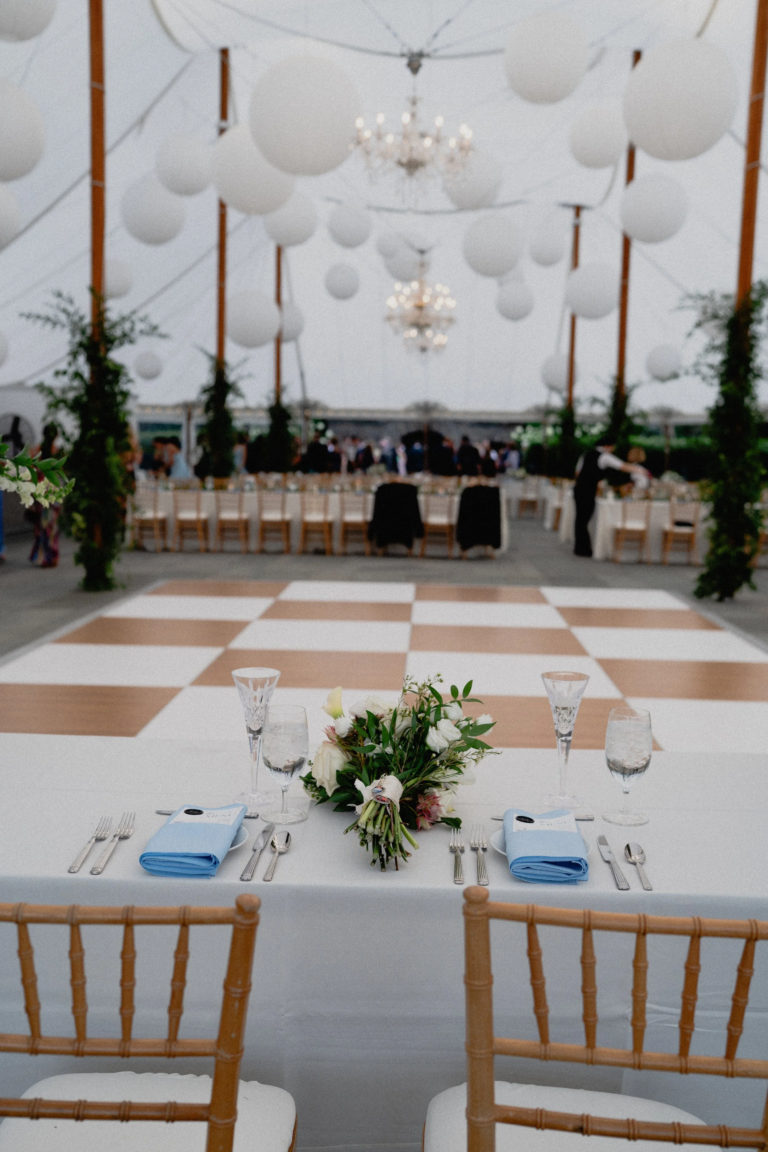 Luxury wedding photography detail of a head table with blush flowers, blue napkins, and glassware beneath an elegant tent.