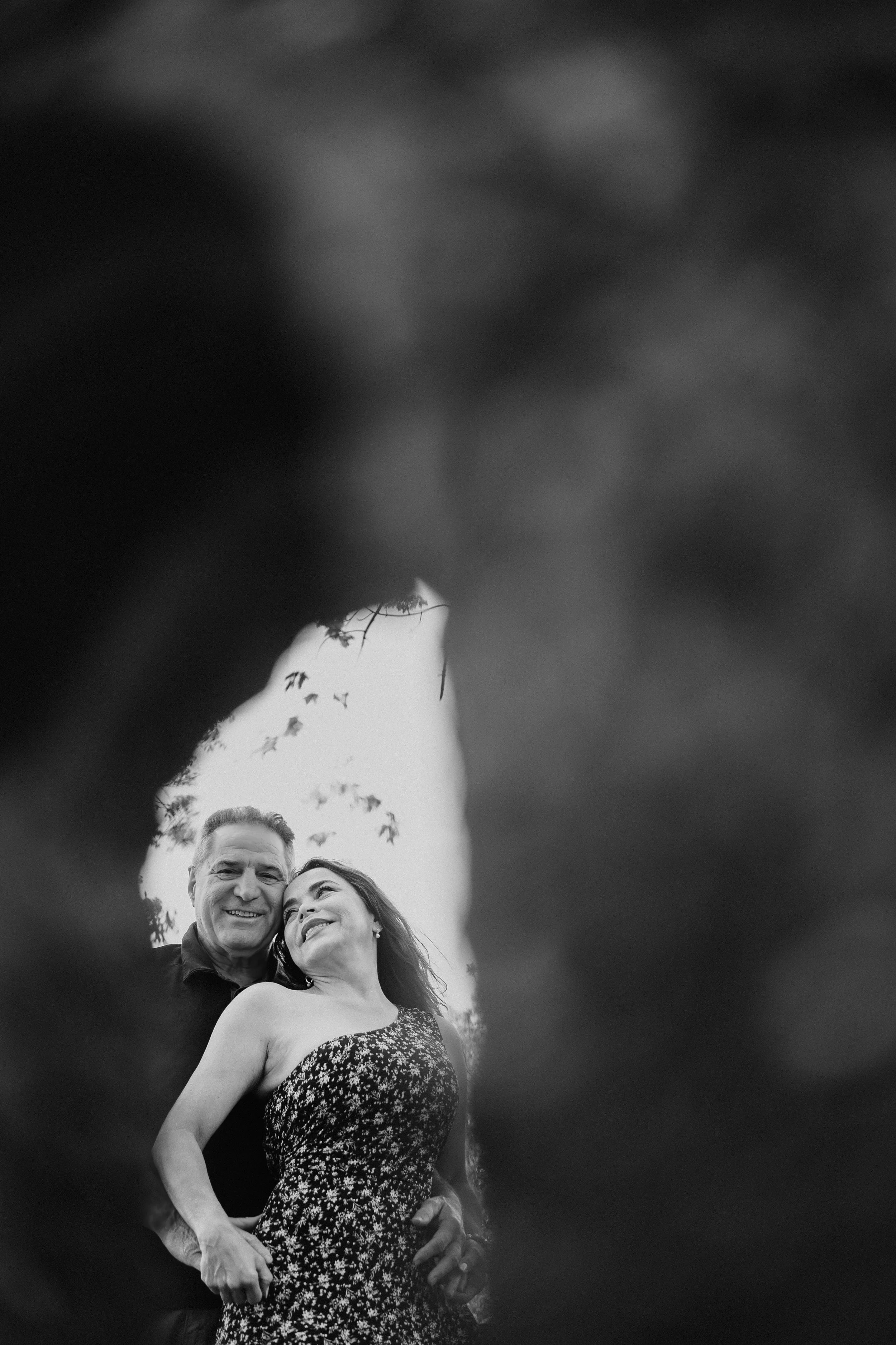NJ Wedding Photography of A black and white photograph of a smiling older man and a woman, seen through a hole or opening in foliage, with trees and sky in the background.