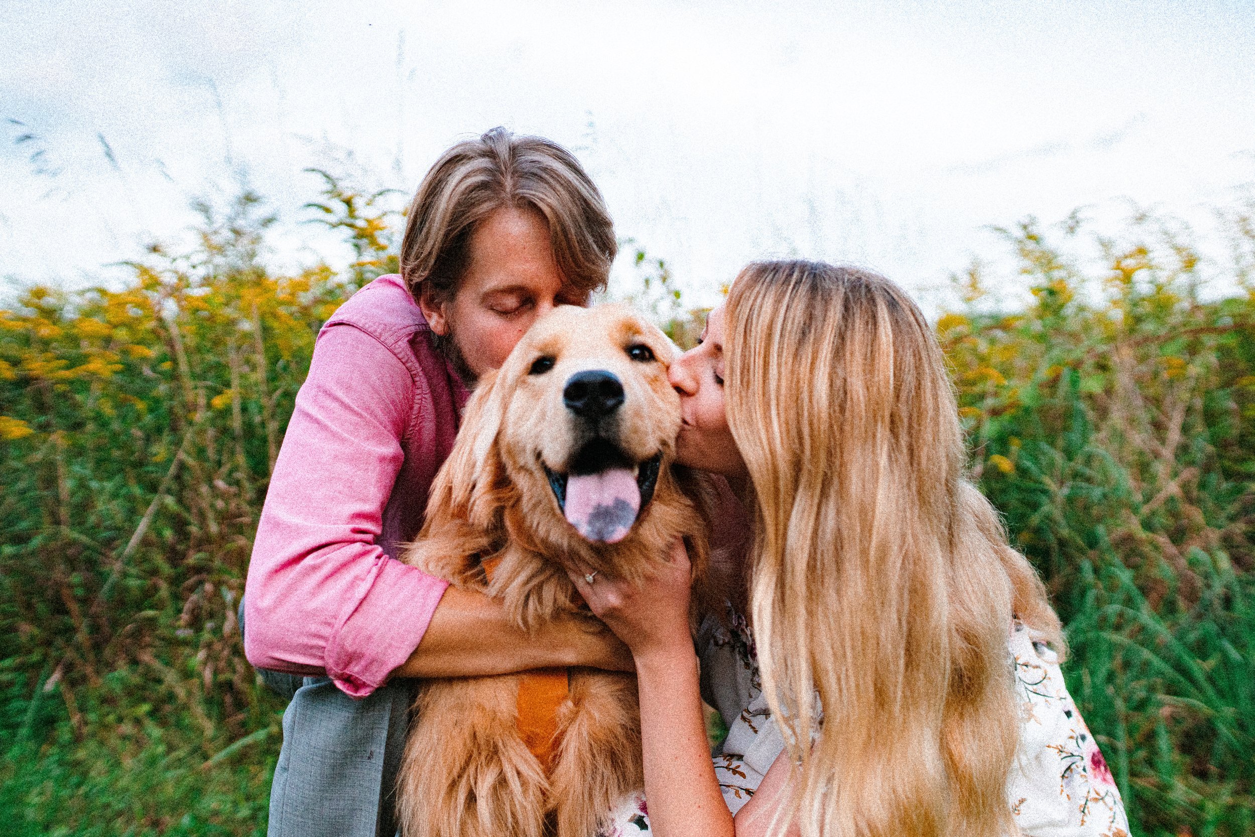Two women and a golden retriever dog outdoors in a field of yellow flowers, with one woman kissing the dog and the other hugging it.